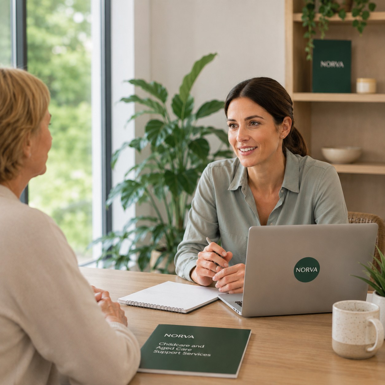 Two women having a conversation at a table in an office. One woman has dark hair and is smiling, with a laptop and notebook in front of her. The other woman has blonde hair and is listening. There are green plants and shelves in the background.