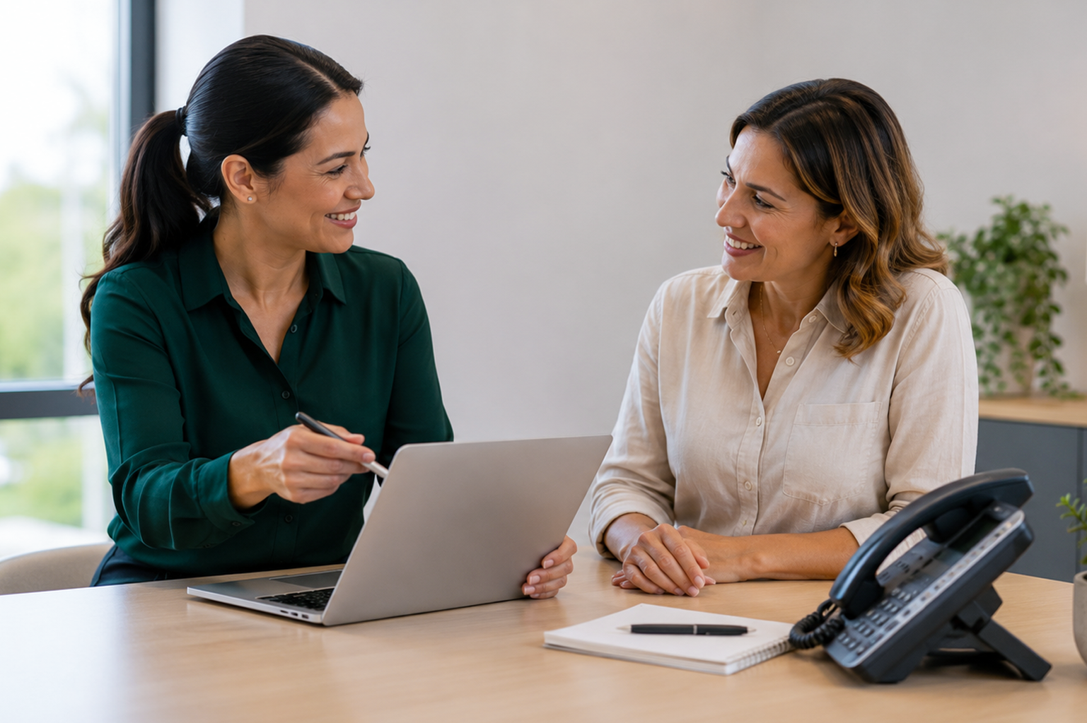 Two women sitting at a desk, having a discussion, with a laptop, phone, notepad, and pen on the desk, in an office with a window and a plant in the background.