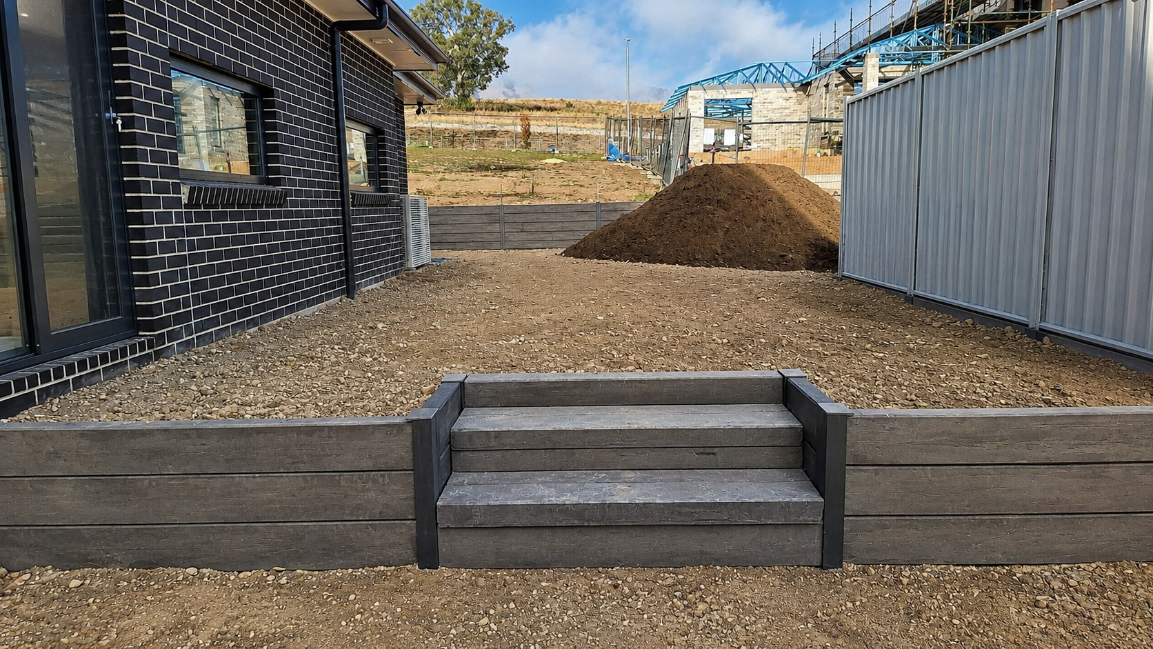 Backyard under construction with steps leading to a dirt yard, a black brick house on the left, a tall metal fence on the right, a mound of dirt in the middle, and a construction site with scaffolding and a partially built structure in the background.