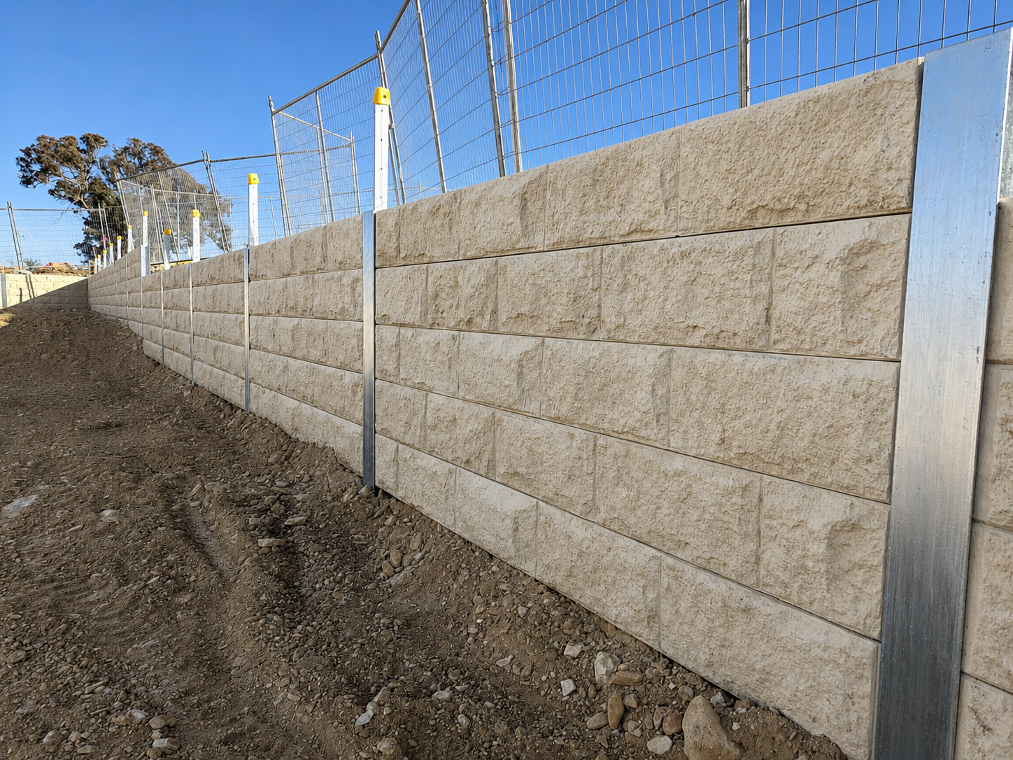 Constructed stone retaining wall with metal fence at the top, on a dirt slope, under a clear blue sky.