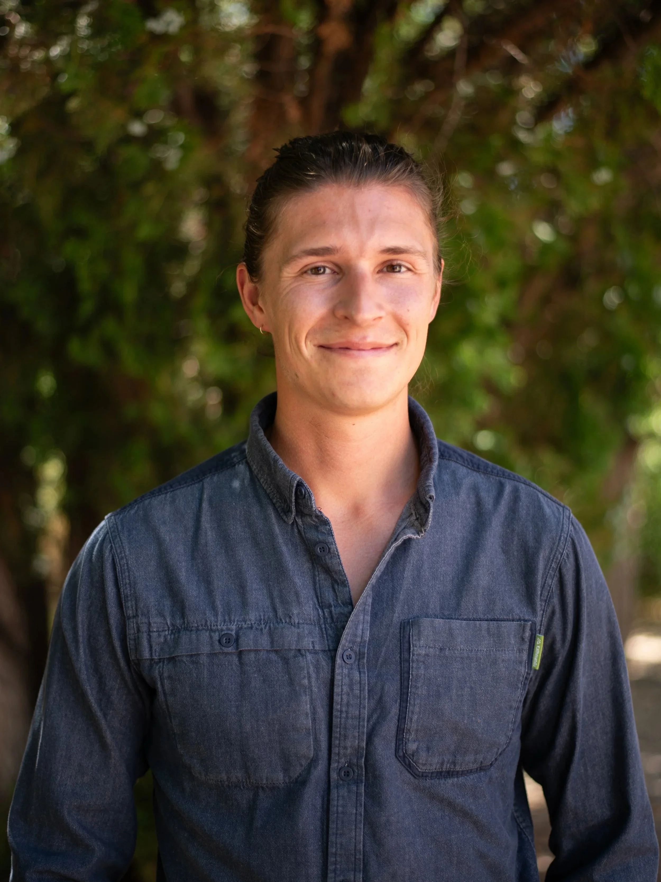 Diego Velasquez with long dark hair tied back, wearing a denim button-up shirt, standing outdoors with blurred green foliage in the background.