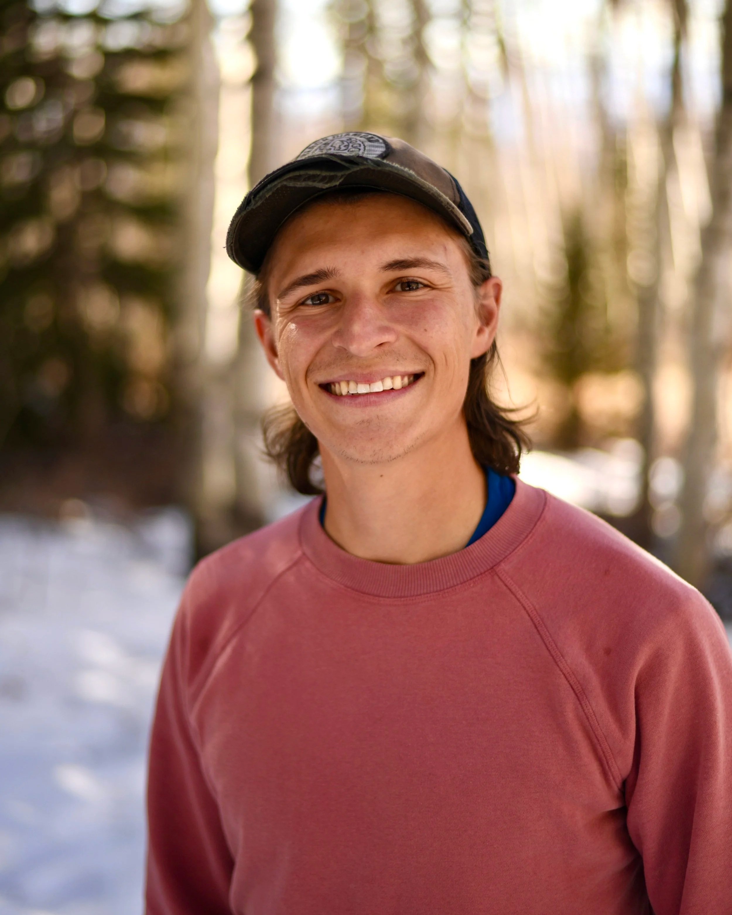 Close-up of a smiling young man wearing a gray cap and a red athletic shirt, outdoors in a forest with sunlight filtering through the trees.