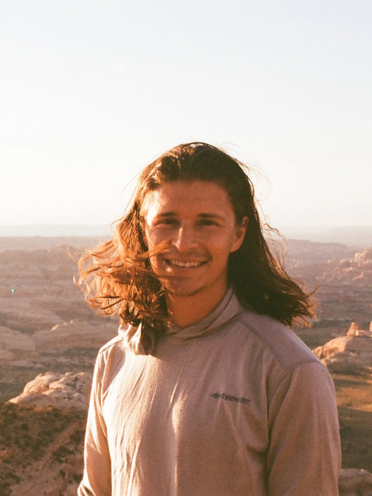 Young man with long hair smiling outdoors during sunset, wearing a light-colored fleece jacket, in a rocky, arid landscape.