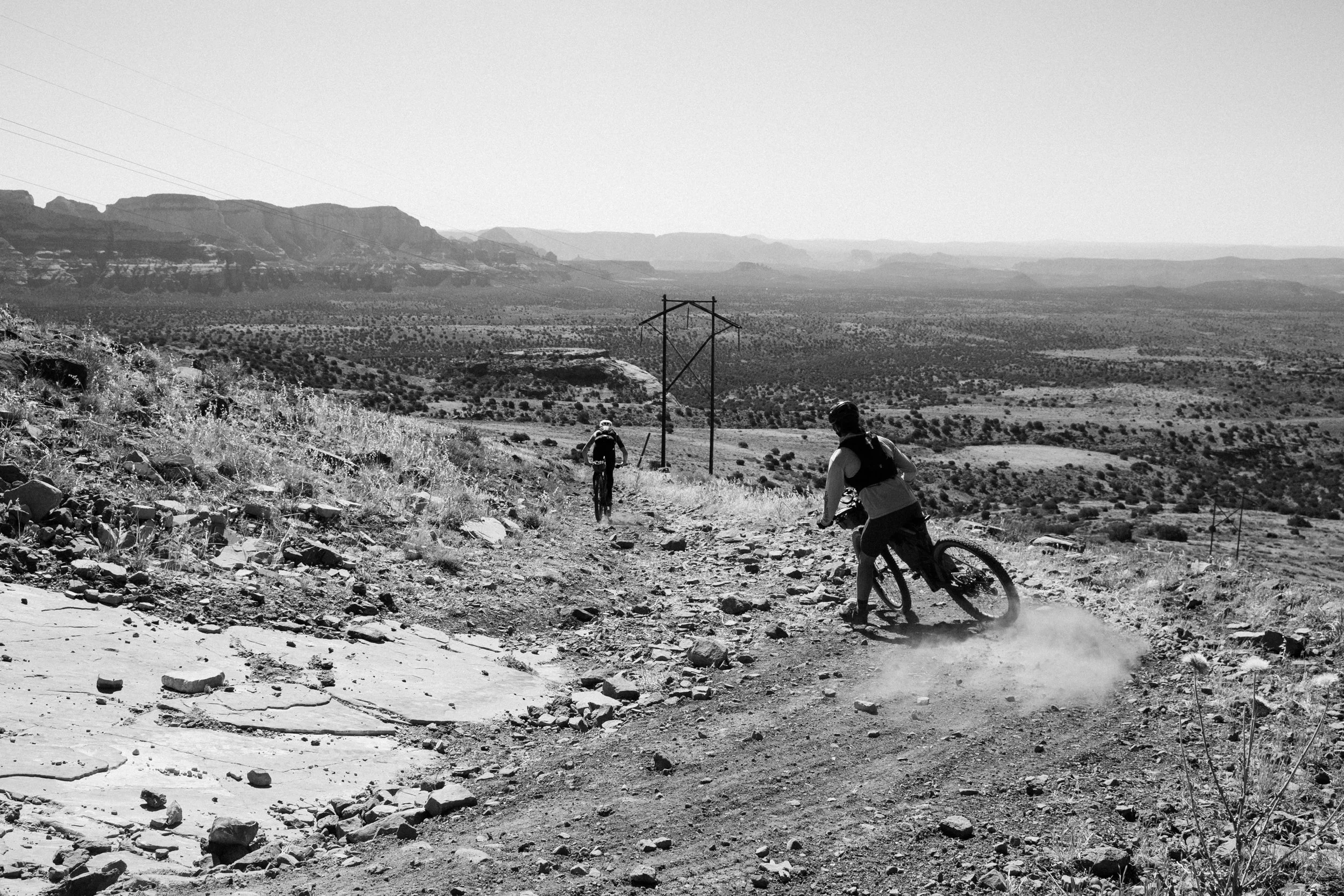 Two mountain bikers riding on a rocky dirt trail in an arid, mountainous landscape with sparse vegetation, power lines, and distant mountains.