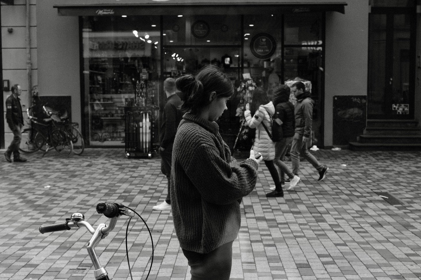 Street photography in bianco e nero: ragazza ferma in strada a Copenaghen che guarda lo smartphone, con manubrio di una bicicletta sfocato in primo piano di Jim Veronese.