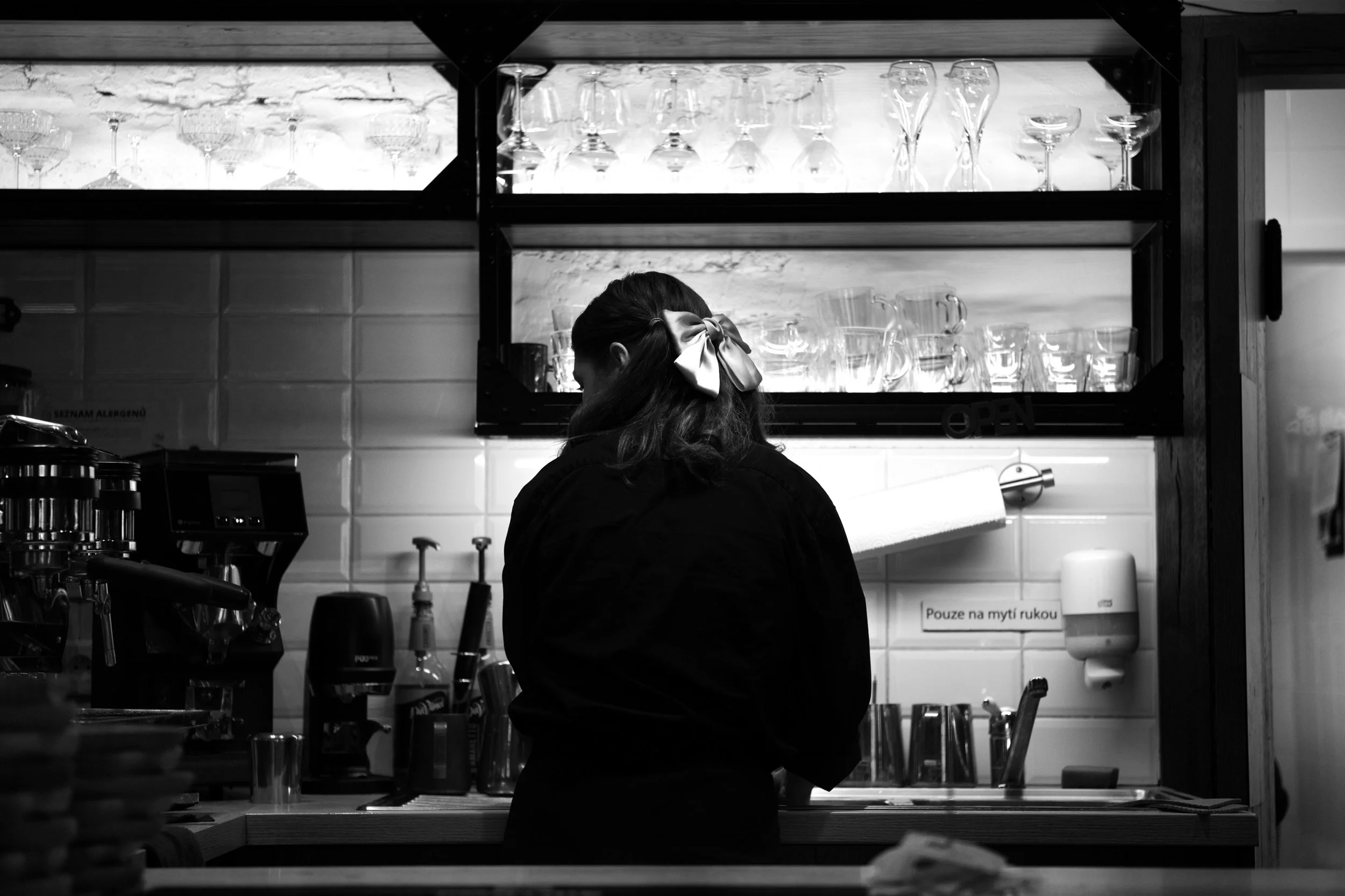 Fotografia in bianco e nero dell'interno di un caffè a Praga. Figura femminile in silhouette contro piastrelle bianche e bicchieri illuminati. Scatto di Jim Veronese.