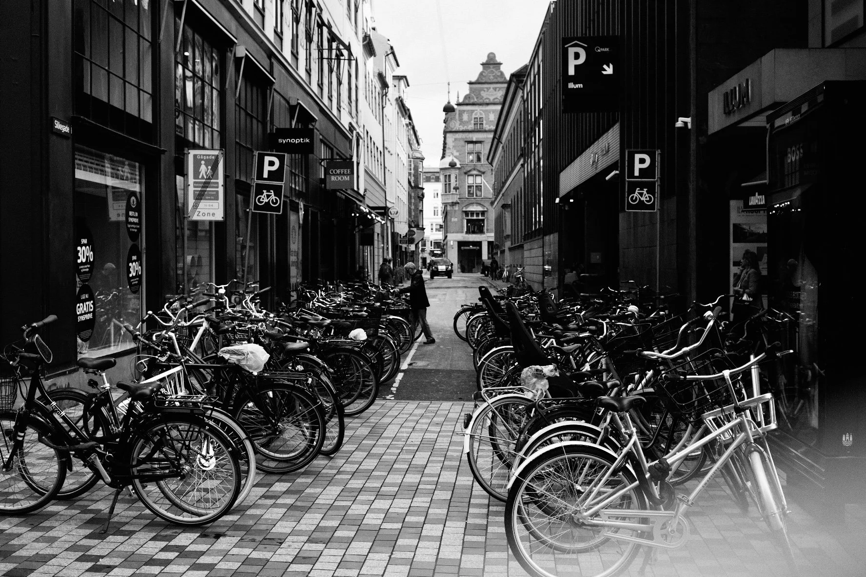 Fotografia in bianco e nero di una strada di Copenaghen densa di biciclette parcheggiate, con linee prospettiche tra i palazzi. Reportage urbano di Jim Veronese.