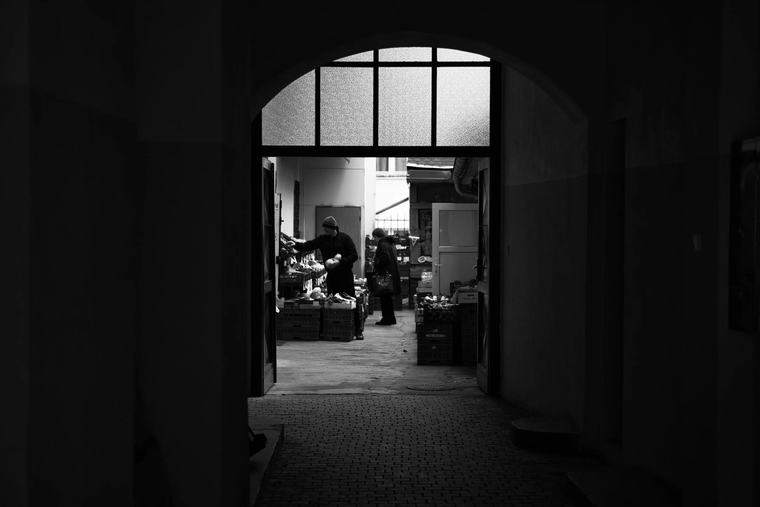 Fotografia in bianco e nero di un profondo arco scuro a Praga che incornicia una scena in un cortile illuminato. Street photography di Jim Veronese.