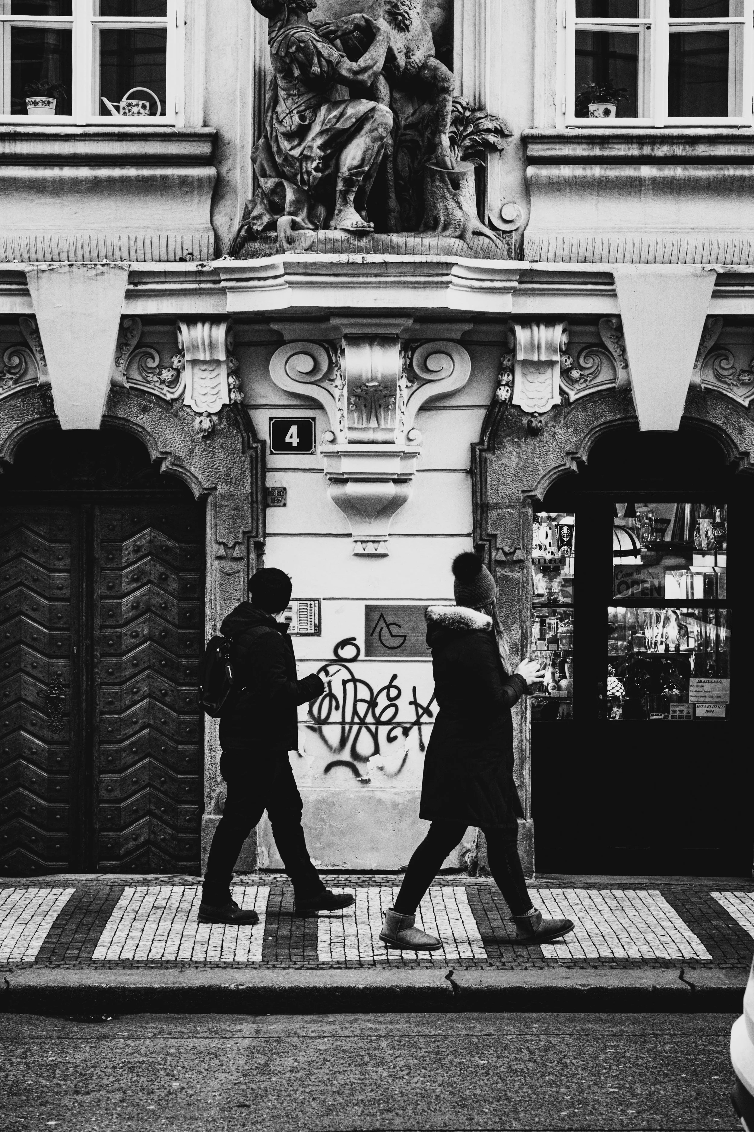 Street photography a Praga: due persone camminano davanti a un palazzo storico con statue classiche, portoni antichi e graffiti. Bianco e nero ad alto contrasto di Jim Veronese.