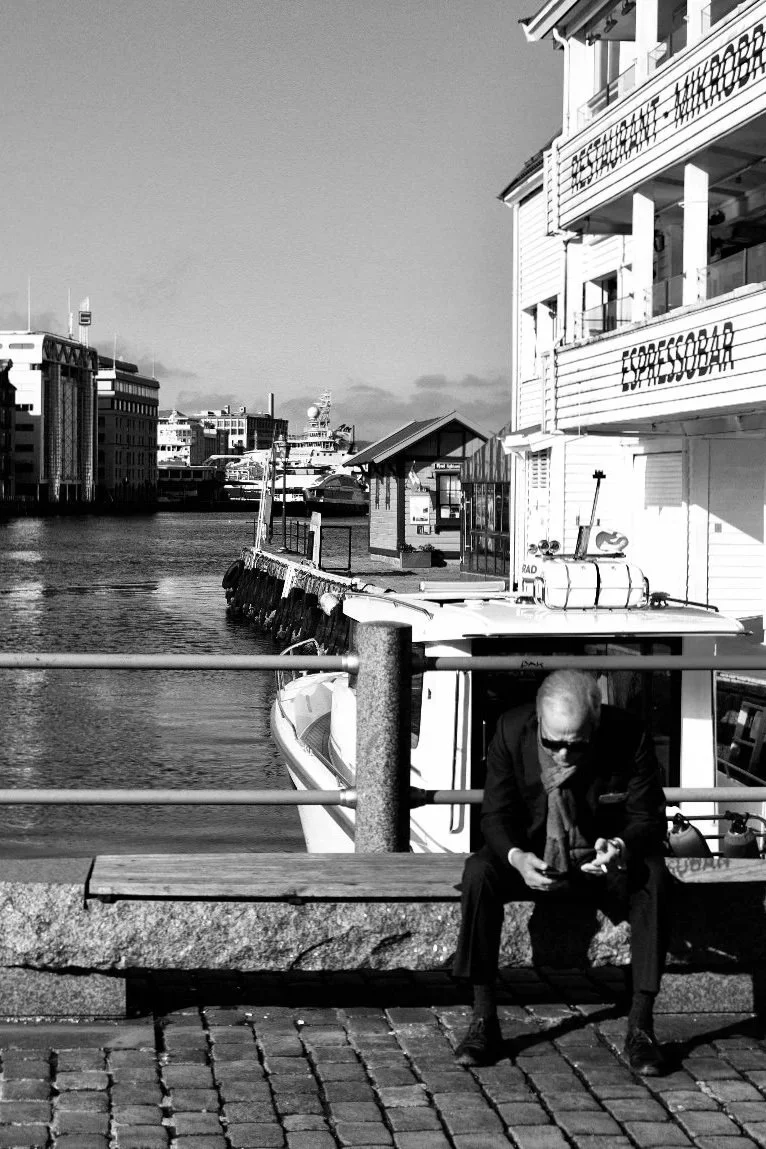 Uomo solitario seduto al porto di Bergen davanti all'Espressobar, street photography in bianco e nero ad alto contrasto di Jim Veronese.