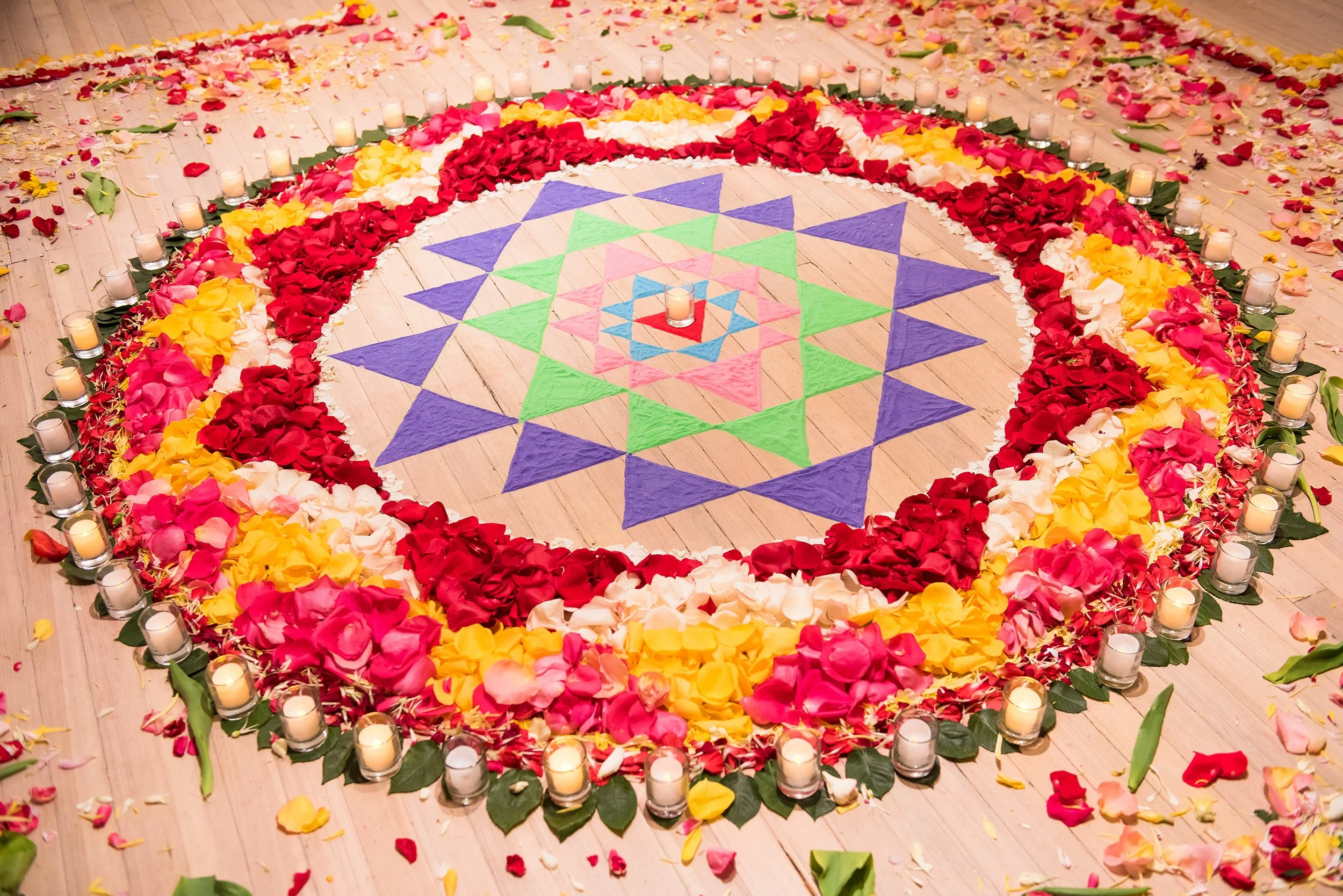 A colorful flower arrangement and petals on the floor forming a geometric pattern, surrounded by candles in glass holders.