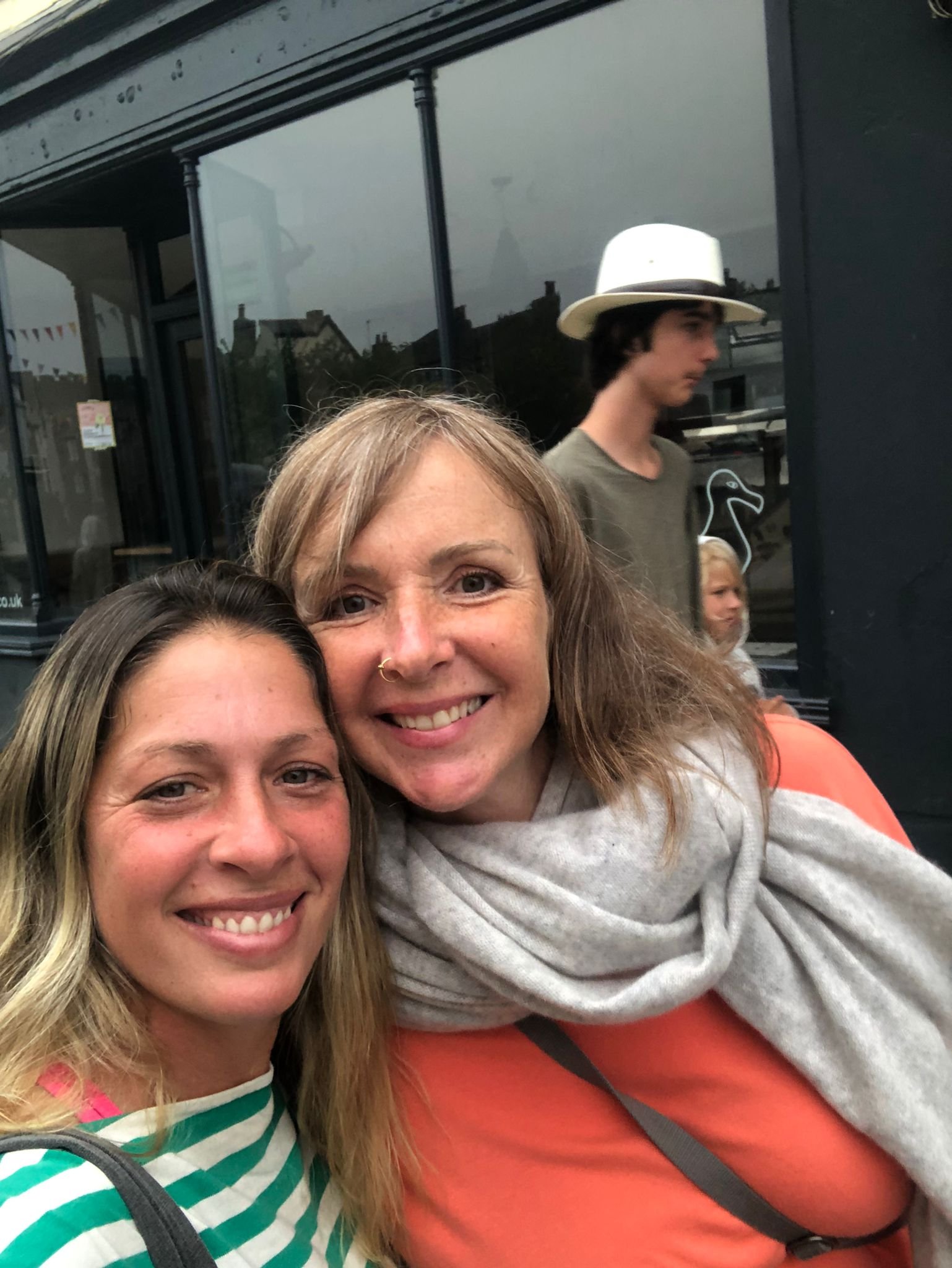 Close-up of Sarah Yantra and her friend Michie taking a selfie in Totnes, Devon,  with a young man in a hat and a utility building reflected in the window behind them.