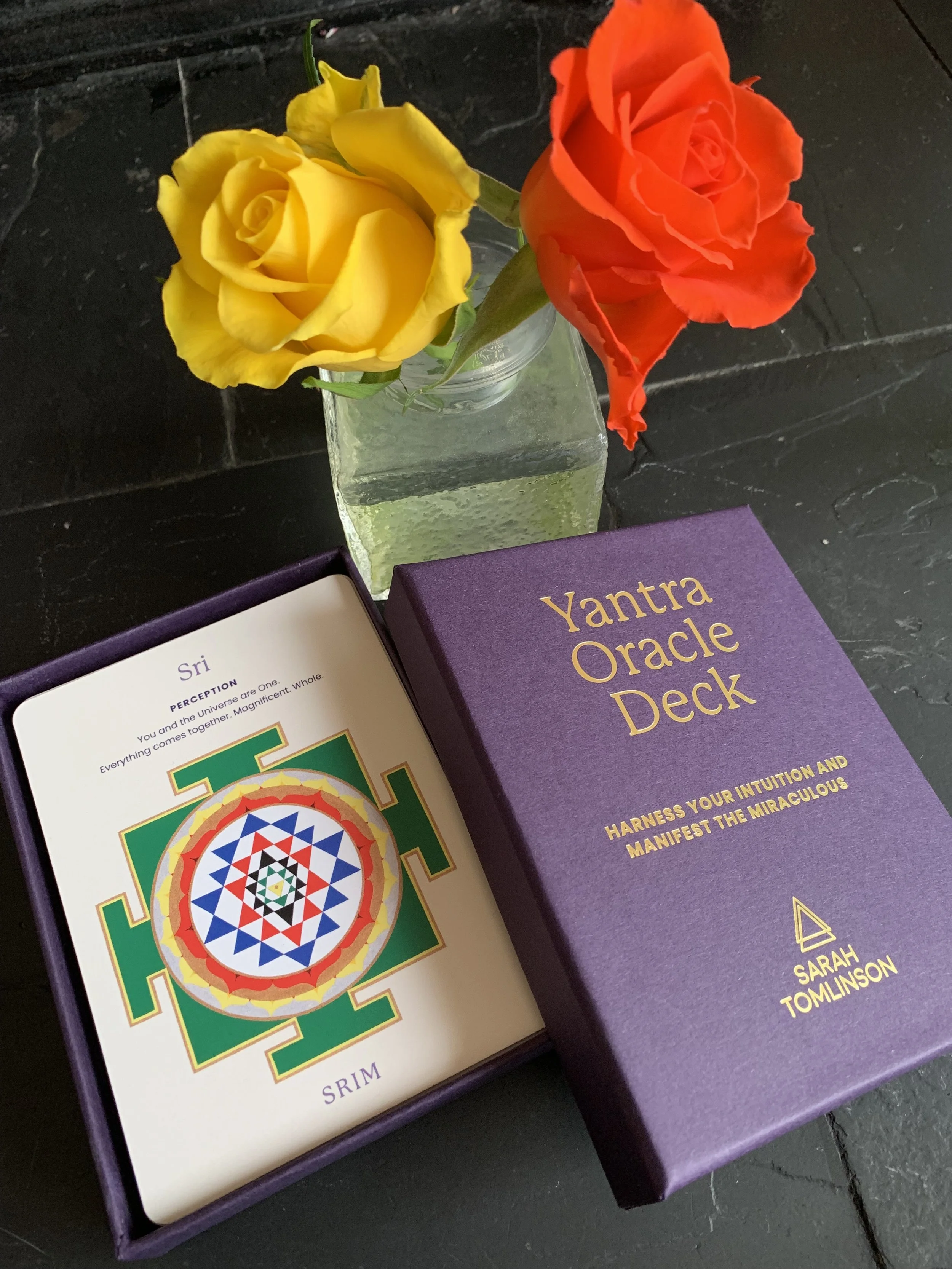 A purple box labeled 'Yantra Oracle Deck', an open card deck with colorful geometric symbols, a yellow rose, an orange rose in a glass vase, and an open book with purple and white pages on a black surface.