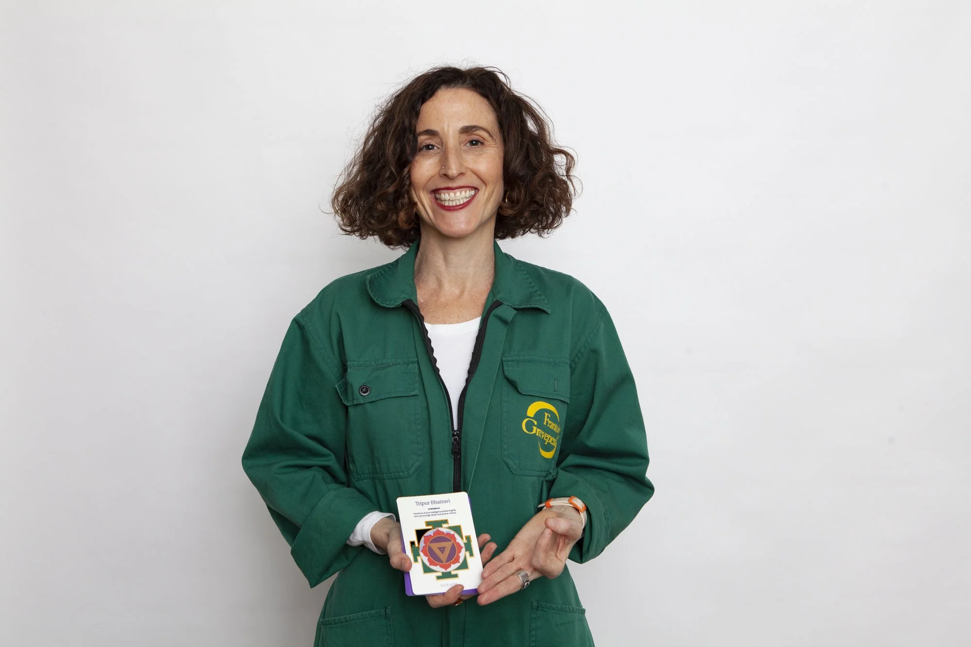 A woman with curly brown hair wearing a green jacket and holding a card. She is smiling and standing in front of a white background.