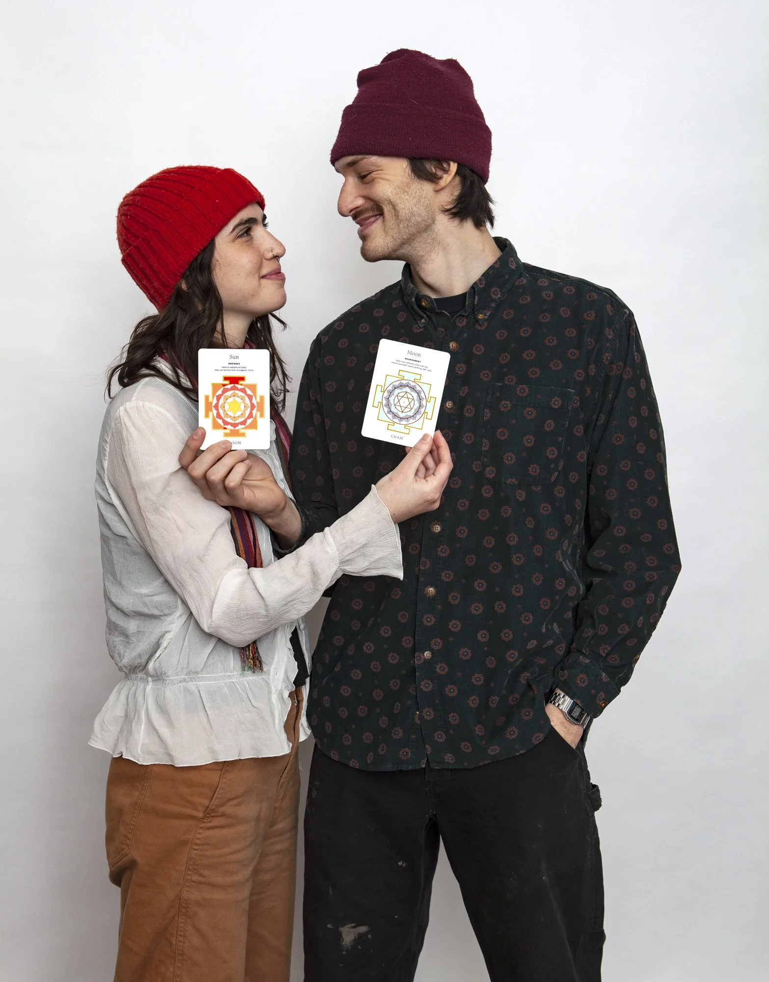 A woman and a man standing close together, facing each other smiling, holding tarot cards with chakra symbols. The woman is wearing a red beanie and a light-colored blouse with brown pants. The man is wearing a maroon beanie and a dark patterned shirt with black pants.