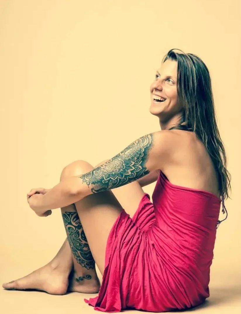 A woman wearing a pink dress sitting on the floor against a plain background, smiling, with tattoos on her arm and leg.