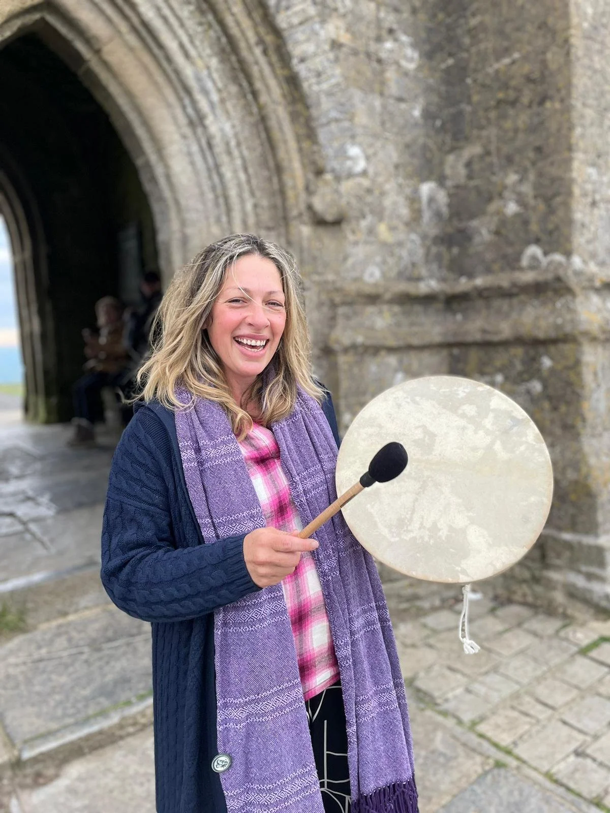 A smiling woman holding a small gong and mallet outside a stone archway, with other people sitting in the background.