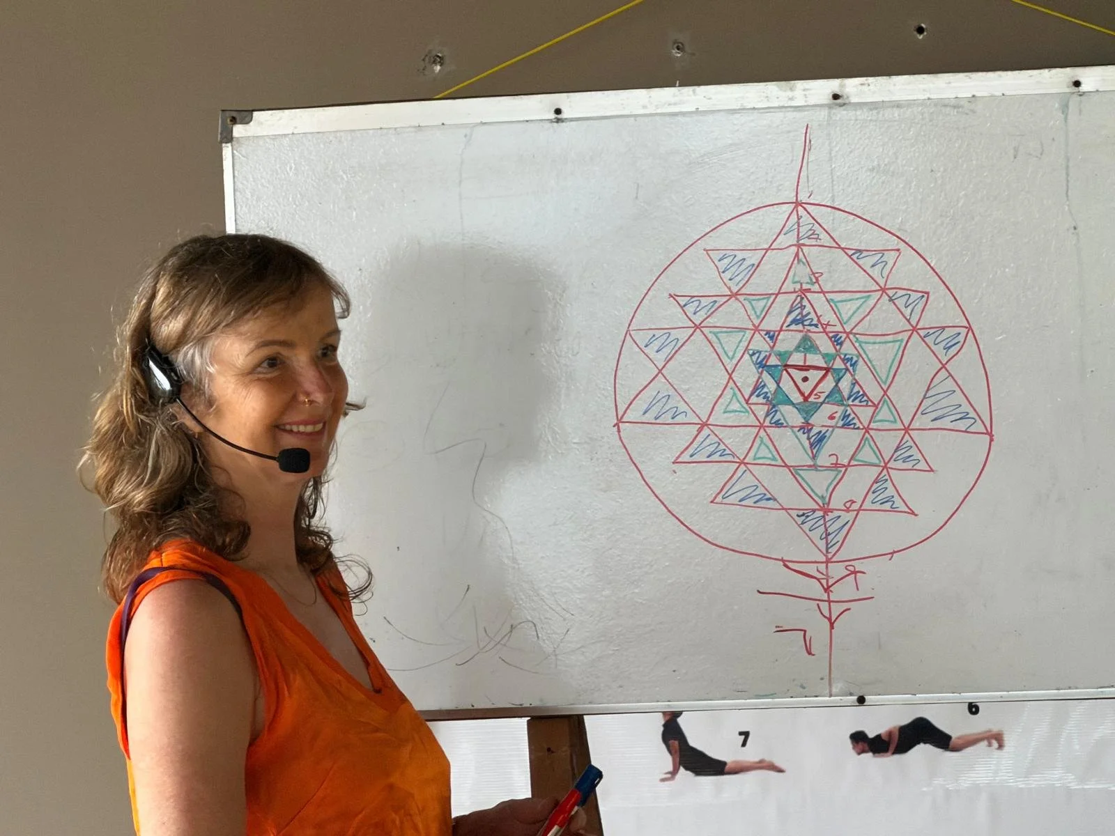 A woman wearing an orange top with a headset, standing in front of a whiteboard with a colorful geometric diagram and illustrations of a person doing yoga poses underneath.