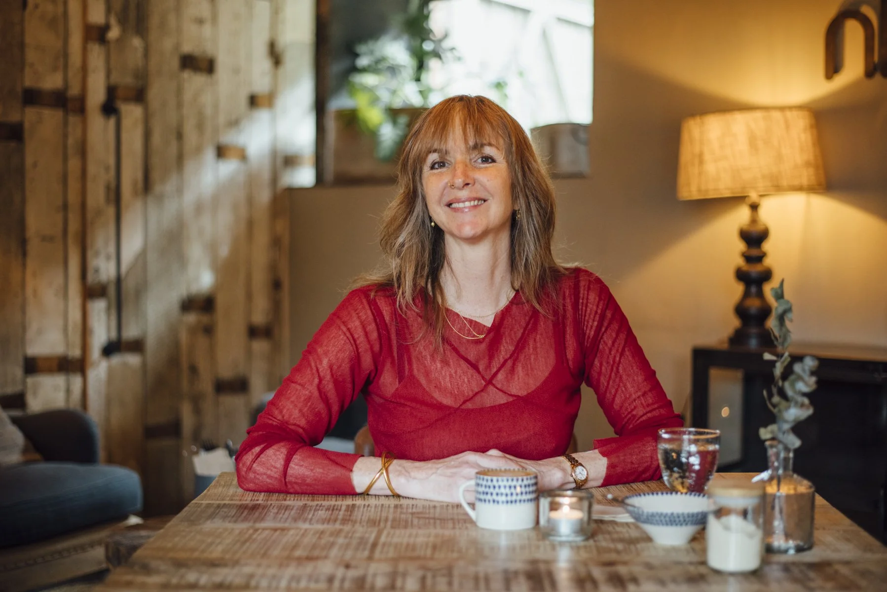 A woman with red hair, wearing a red top, sitting at a wooden table in a cozy room with warm lighting, surrounded by decorative candles and tableware.