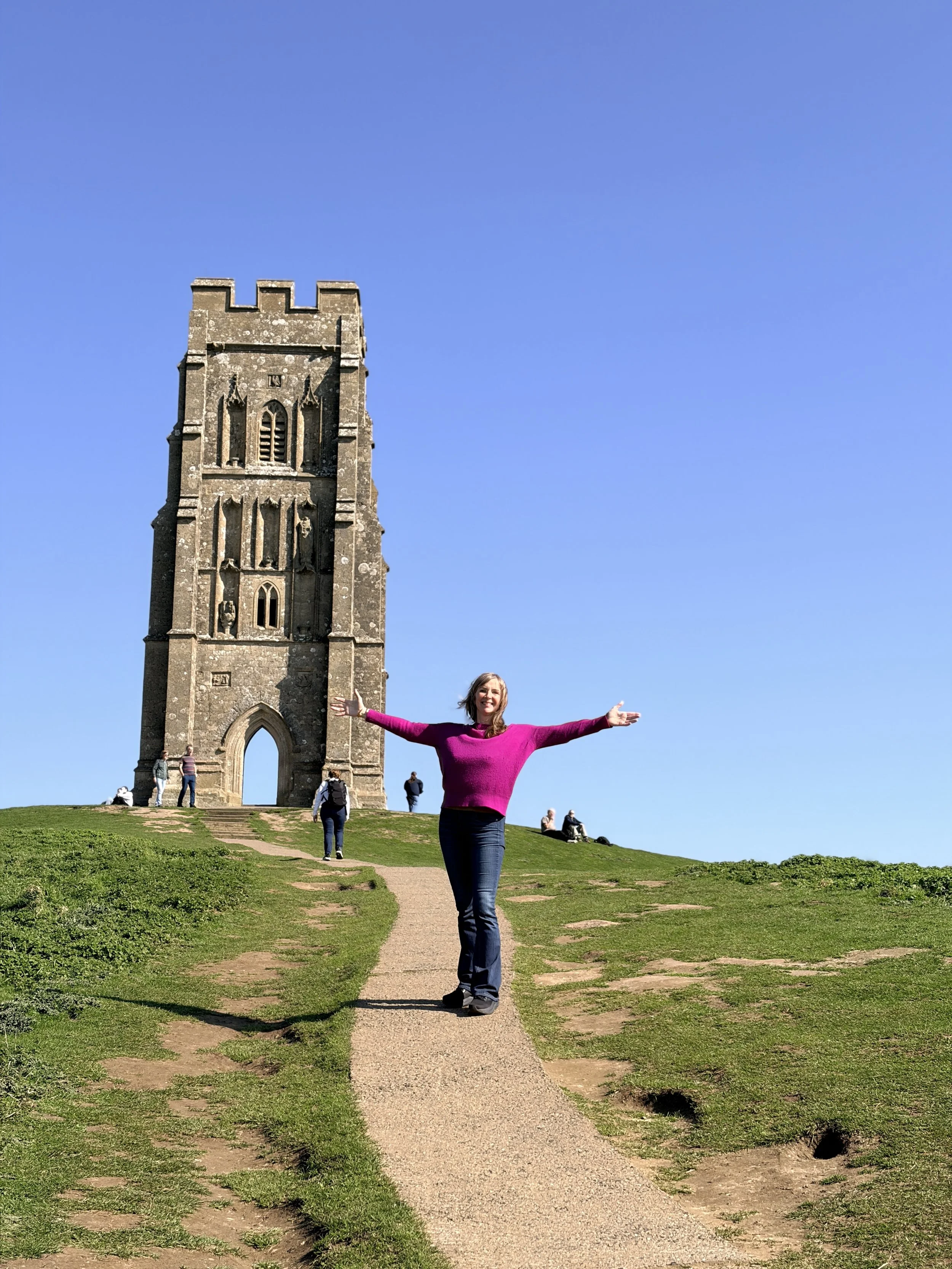 Sarah Yantra in Glastonbury Tor with arms outstretched standing  under a clear blue sky.
