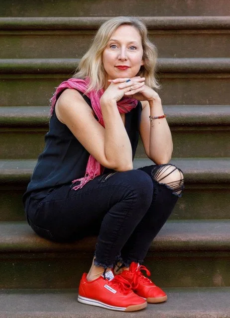 A woman with blonde hair, wearing a black sleeveless top, ripped black jeans, red sneakers, and a pink scarf, sitting on outdoor stairs with her hands resting under her chin and looking at the camera.