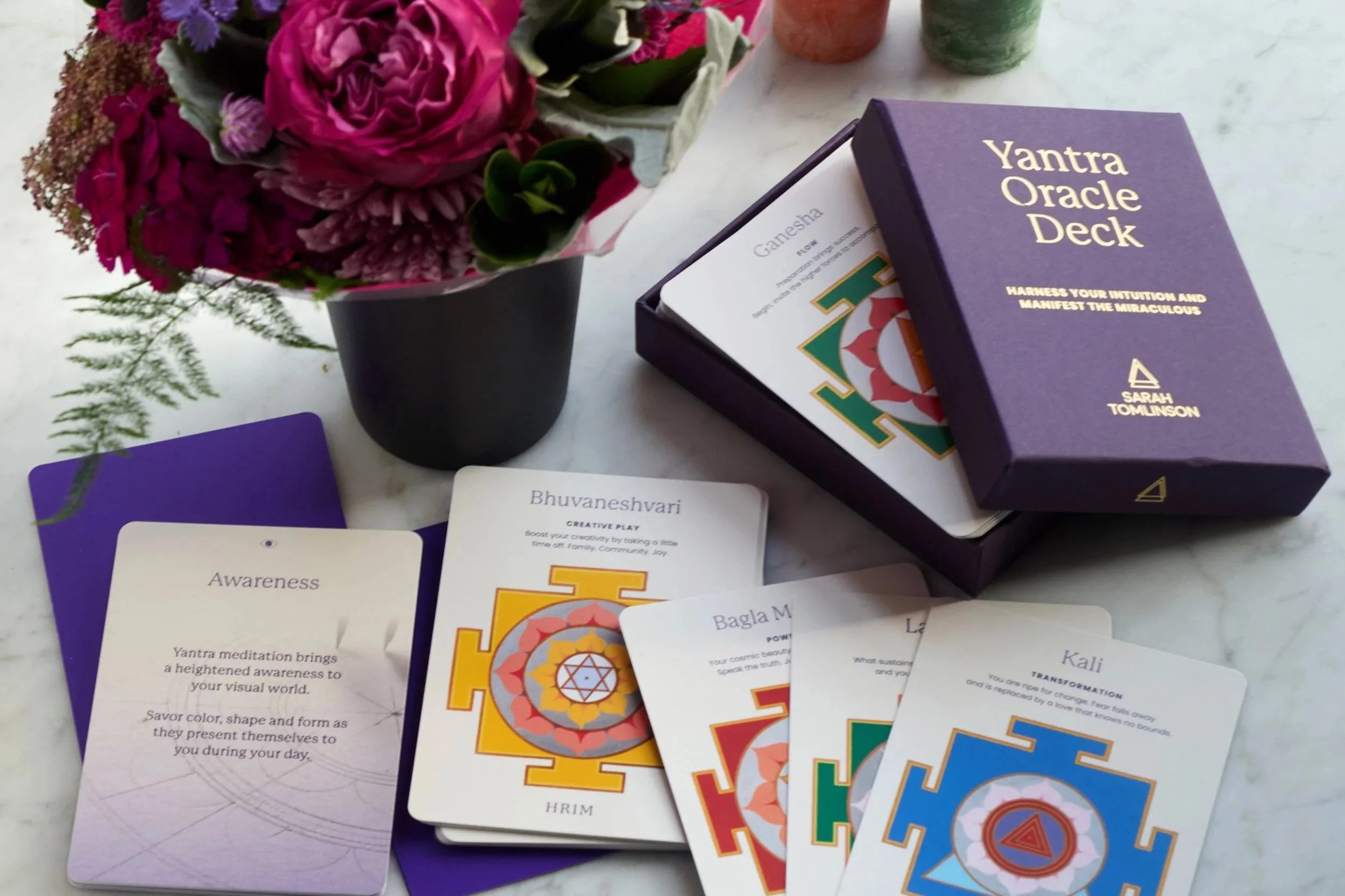 Colorful deck of Oracle cards with the title 'Yantra Oracle Deck' on the box, alongside a small bouquet of pink and purple flowers in a black vase, on a white surface.
