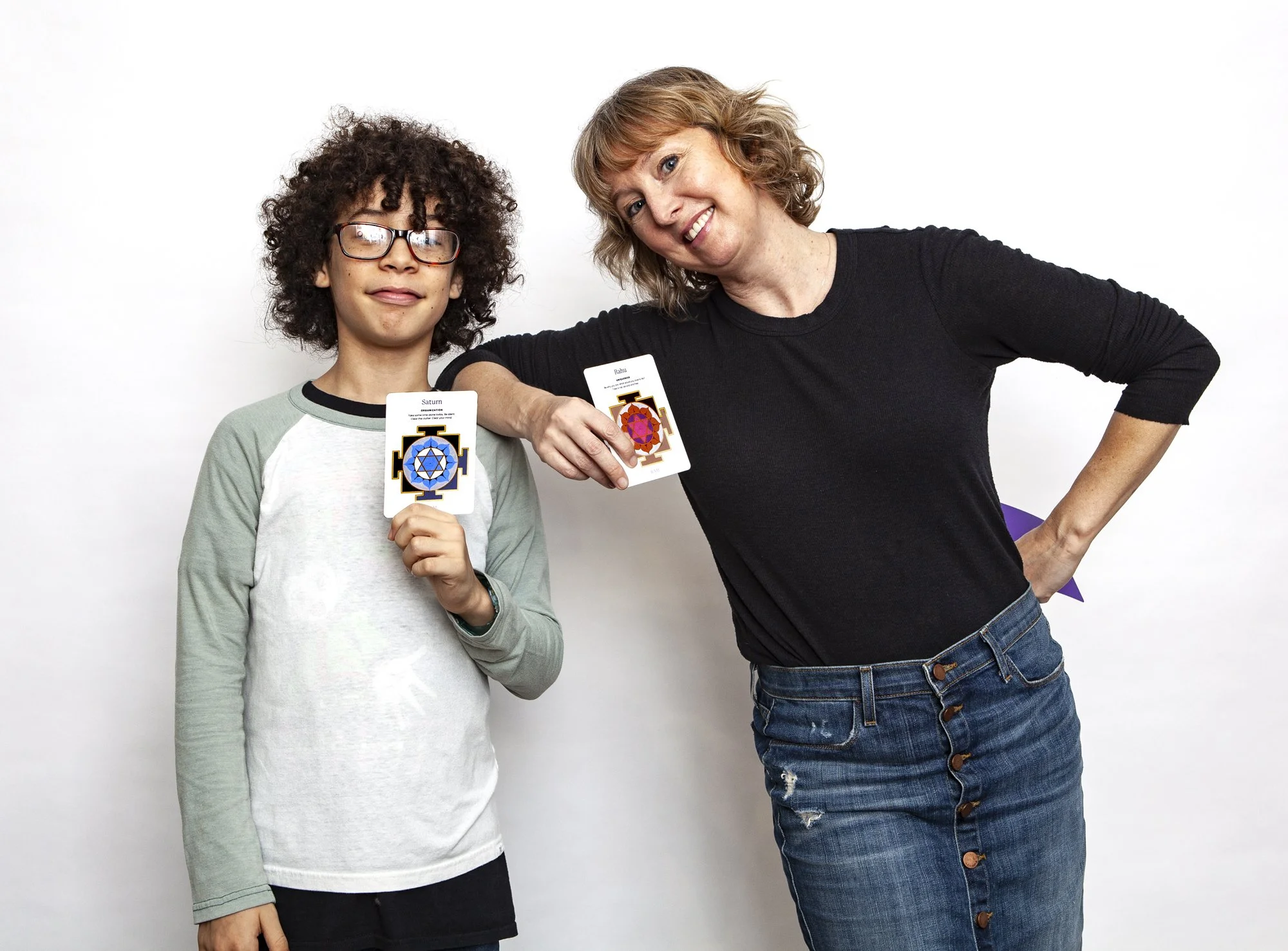 A woman and a boy standing against a white background, smiling. The boy has curly hair, glasses, and wears a light-colored long-sleeve shirt. The woman has shoulder-length wavy hair and wears a black shirt. Both are holding Yantra Oracle Deck cards.
