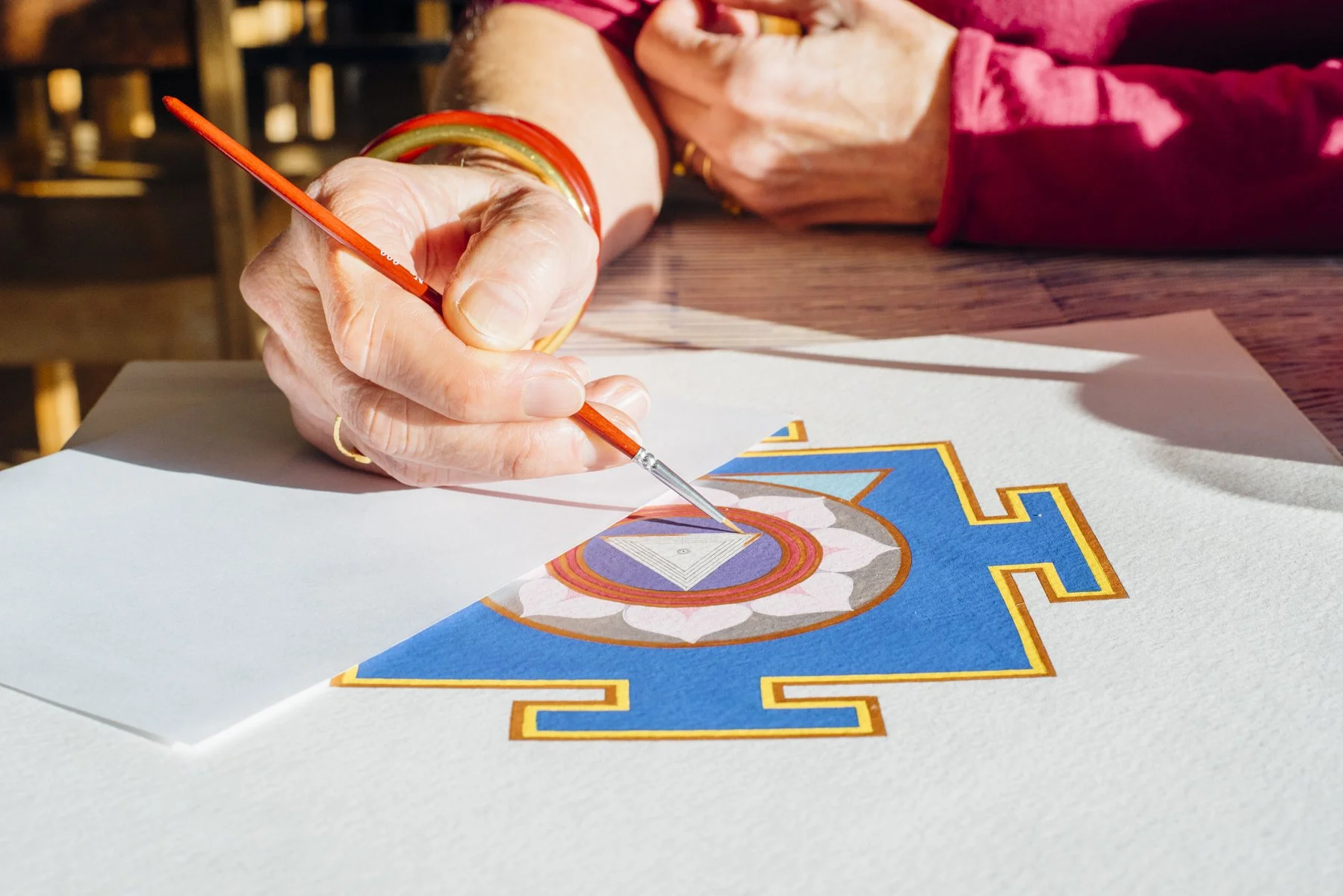 A person drawing a geometric design on a piece of paper with a thin red paintbrush. The paper is on a wooden table, and the person is wearing colorful bangles on their wrist.