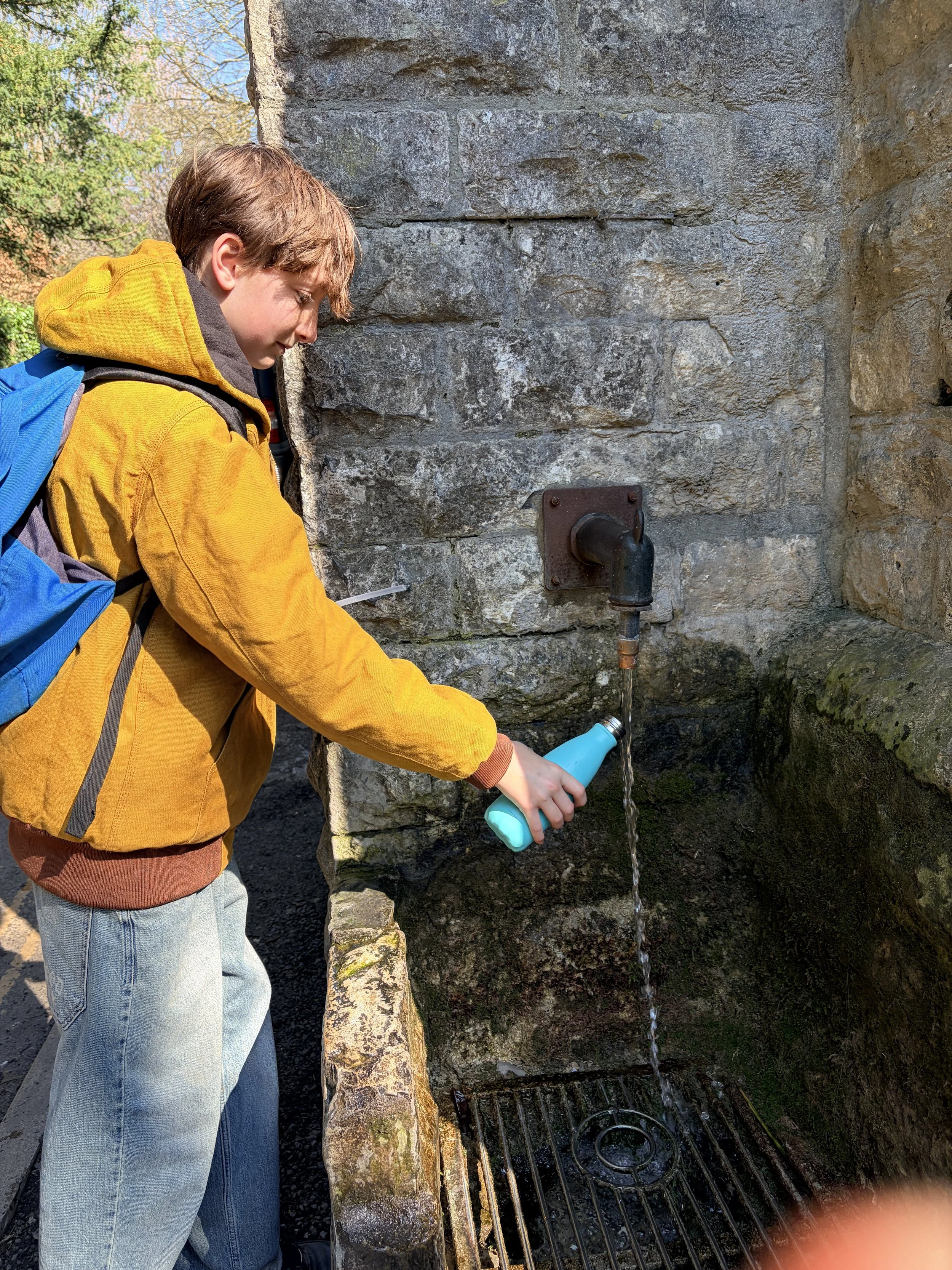 A young person in a yellow jacket and gray jeans is filling a blue water bottle at an outdoor stone water fountain with Spring Water from Glastonbury Tor.
