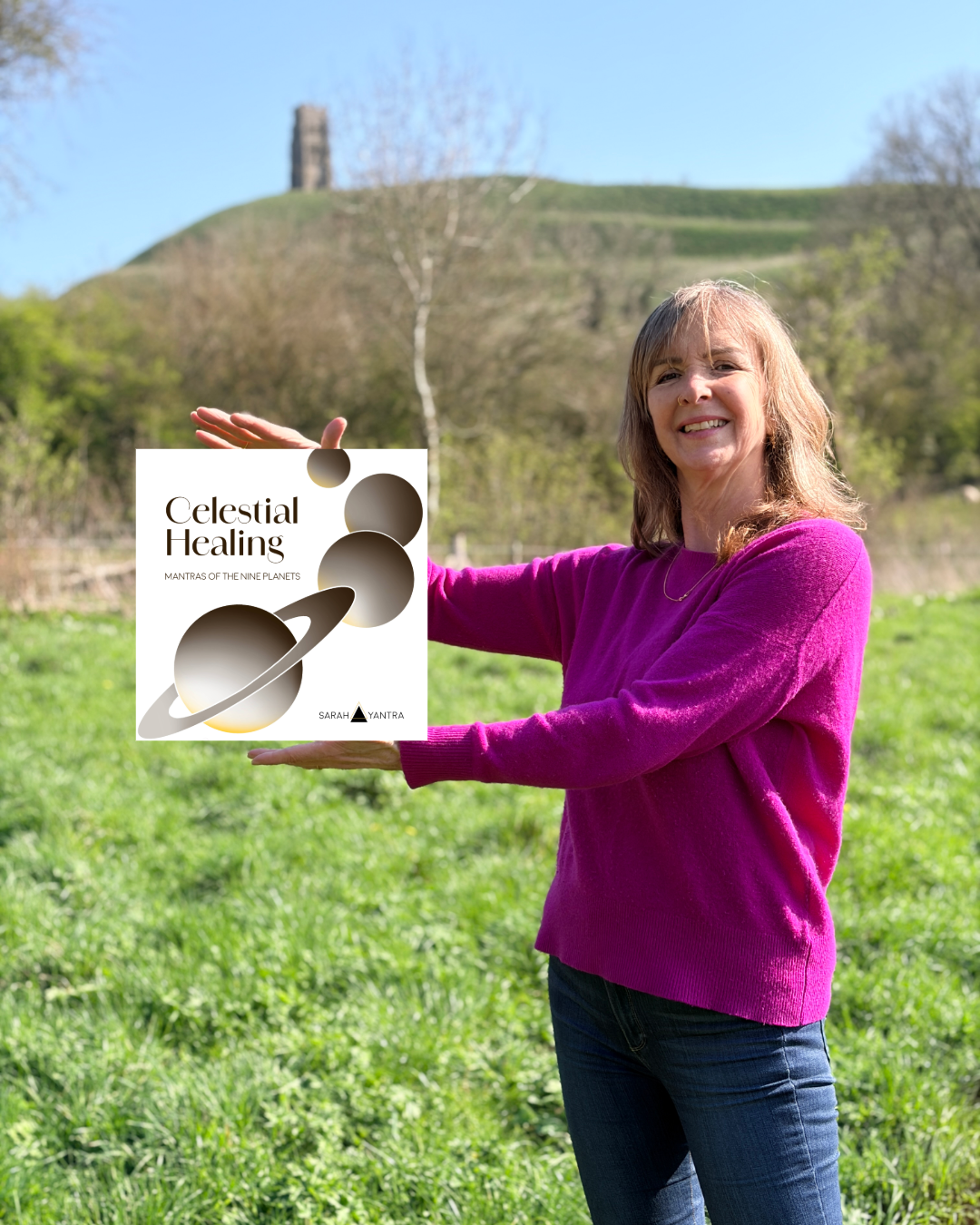 A woman wearing a pink sweater standing outdoors on a grassy field, smiling and holding a book titled 'Celestial Healing' by Sarah Yantra with a background of trees and a hill.