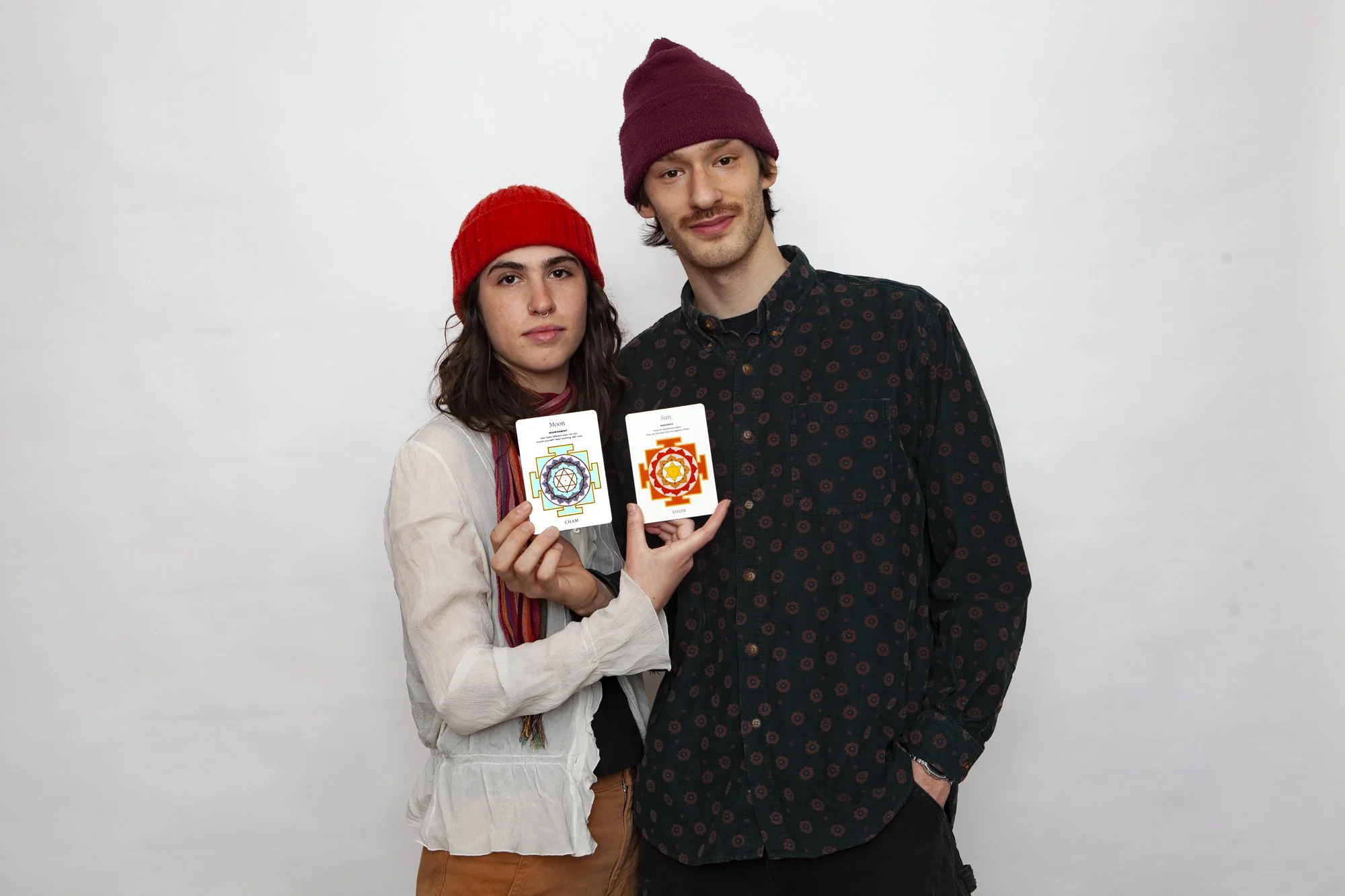 A young woman and a young man stand against a plain white background, holding up two colorful cards with mandala designs and labels 'Moon' and 'Sun'.
