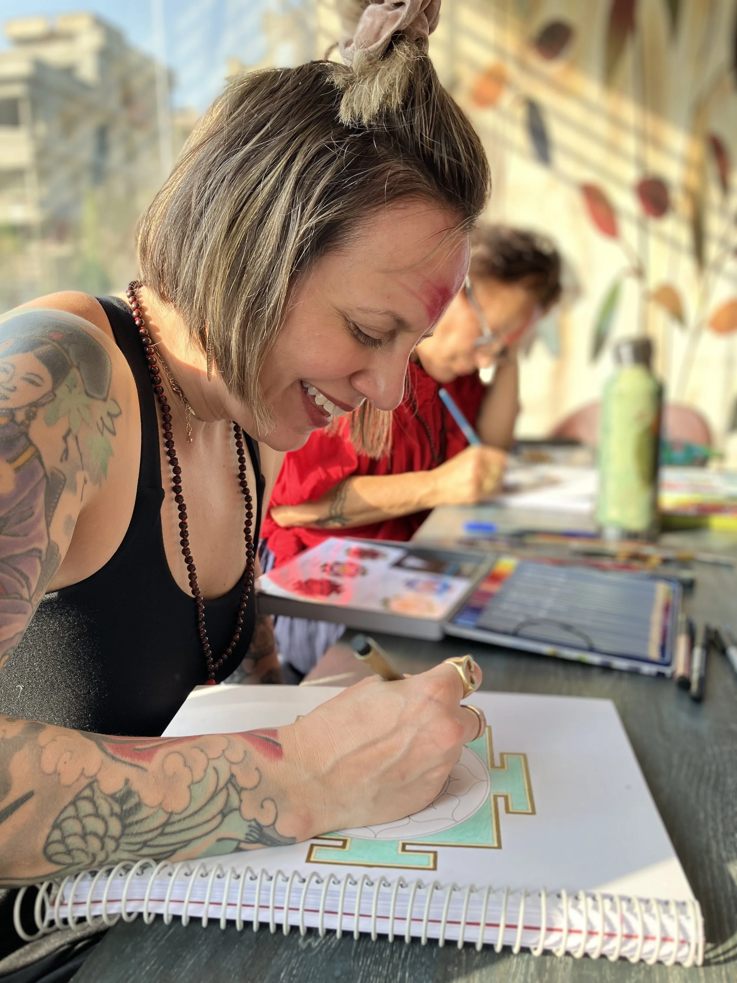 A tattooed woman with short hair and a beaded necklace is coloring in a detailed coloring book at a table. In the background, an older woman is also engaged in coloring. The scene is set indoors with a mural or wall art featuring colorful leaf designs.