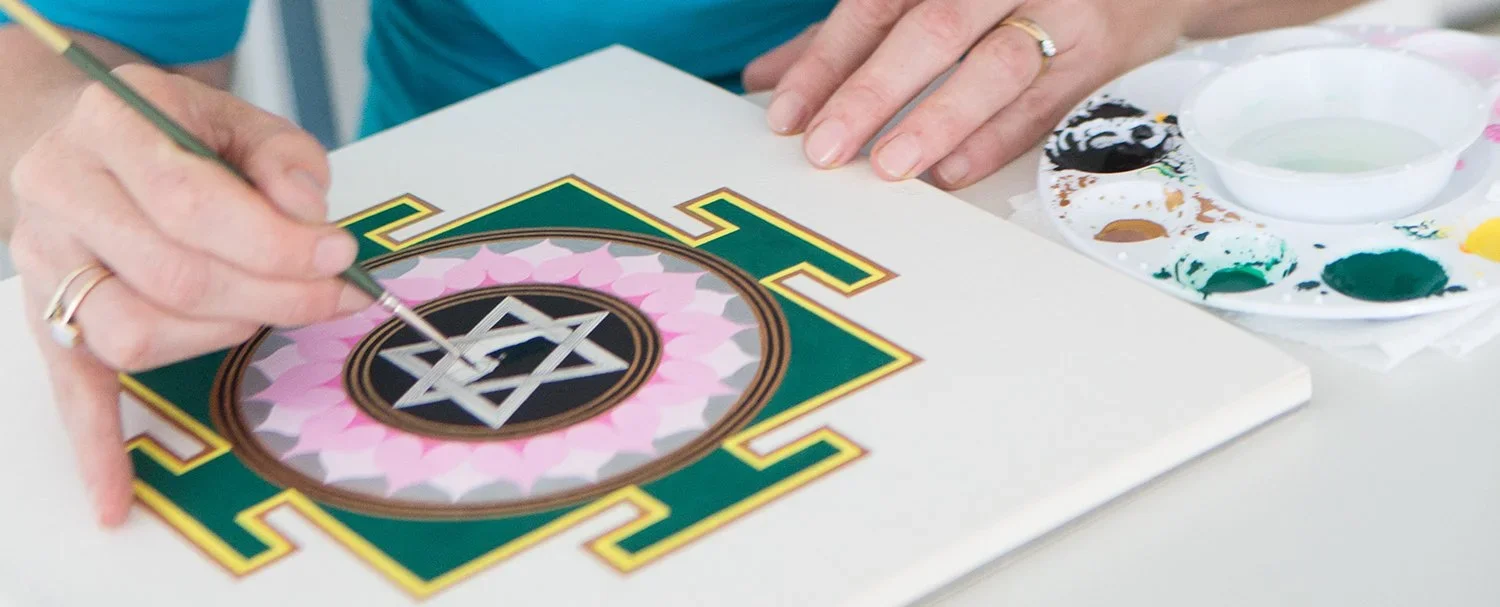 Person painting a geometric design with watercolor paints on canvas, with a palette and water cup nearby.