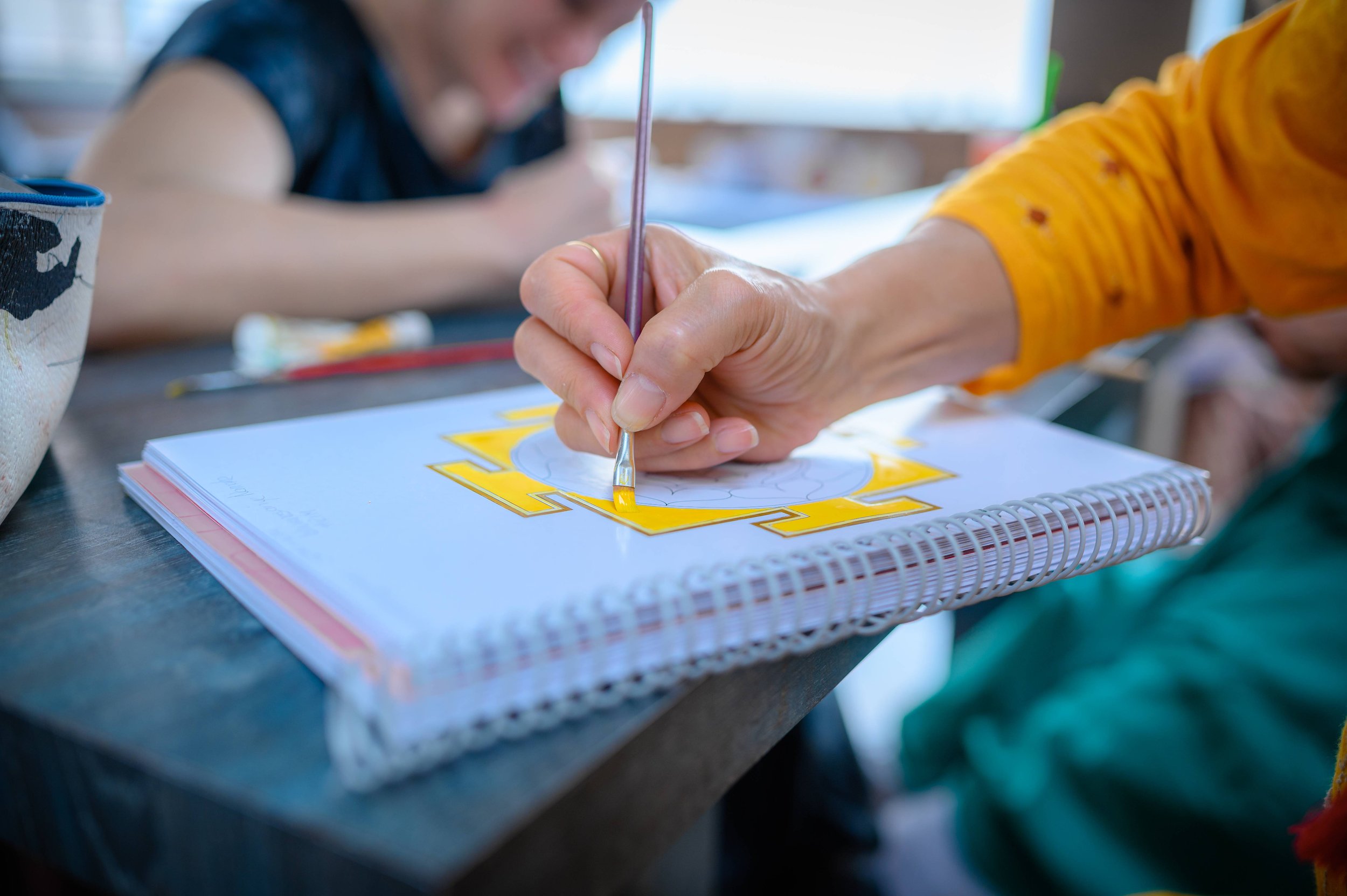Close-up of a person painting a yellow design on a white notebook with a small brush, while another person in the background is smiling and blurred.