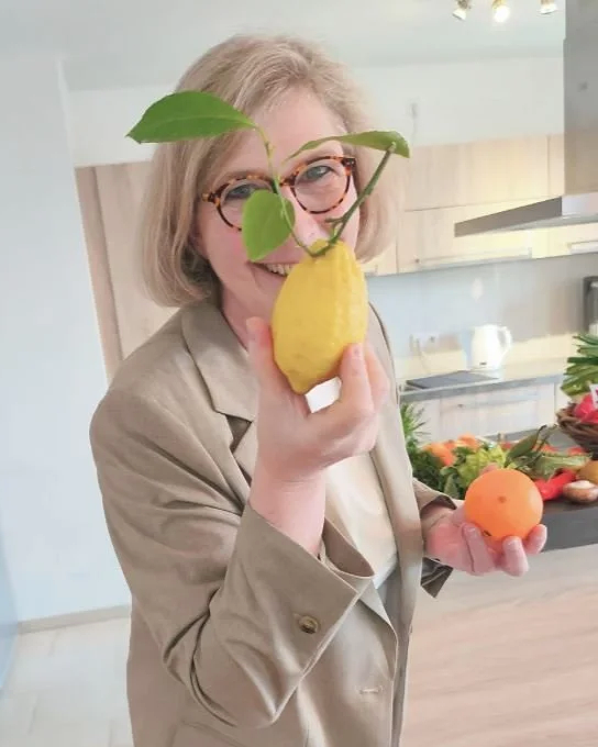 Frau hält einen gelben Birnen mit grünen Blättern und einen kleinen orangenen Ball in der Hand, steht in einer modernen Küche.