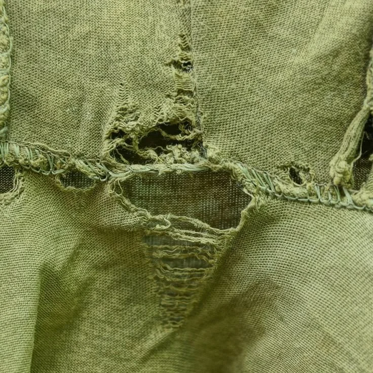 Close-up of a heavily worn and torn piece of olive green fabric, with frayed edges and visible stitching.