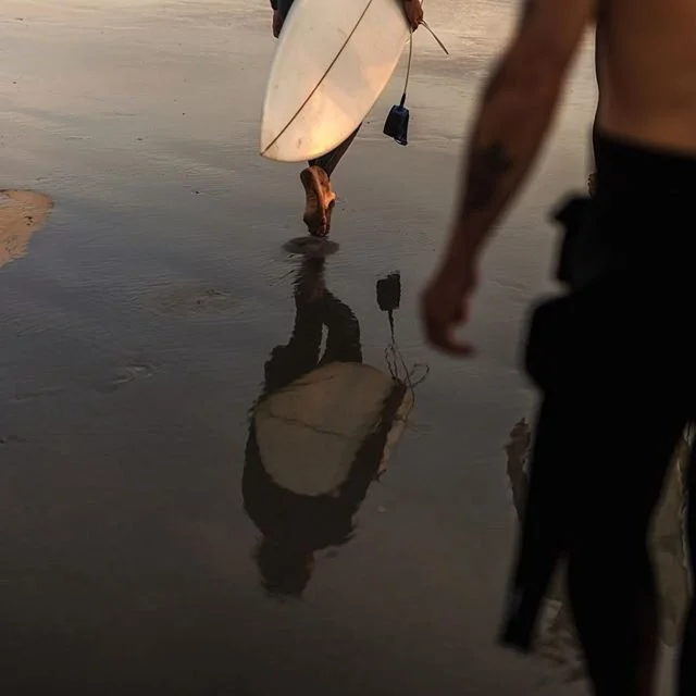 Two people walking on a wet beach, one carrying a surfboard and the other holding a camera, with their reflections visible on the wet sand at sunset.