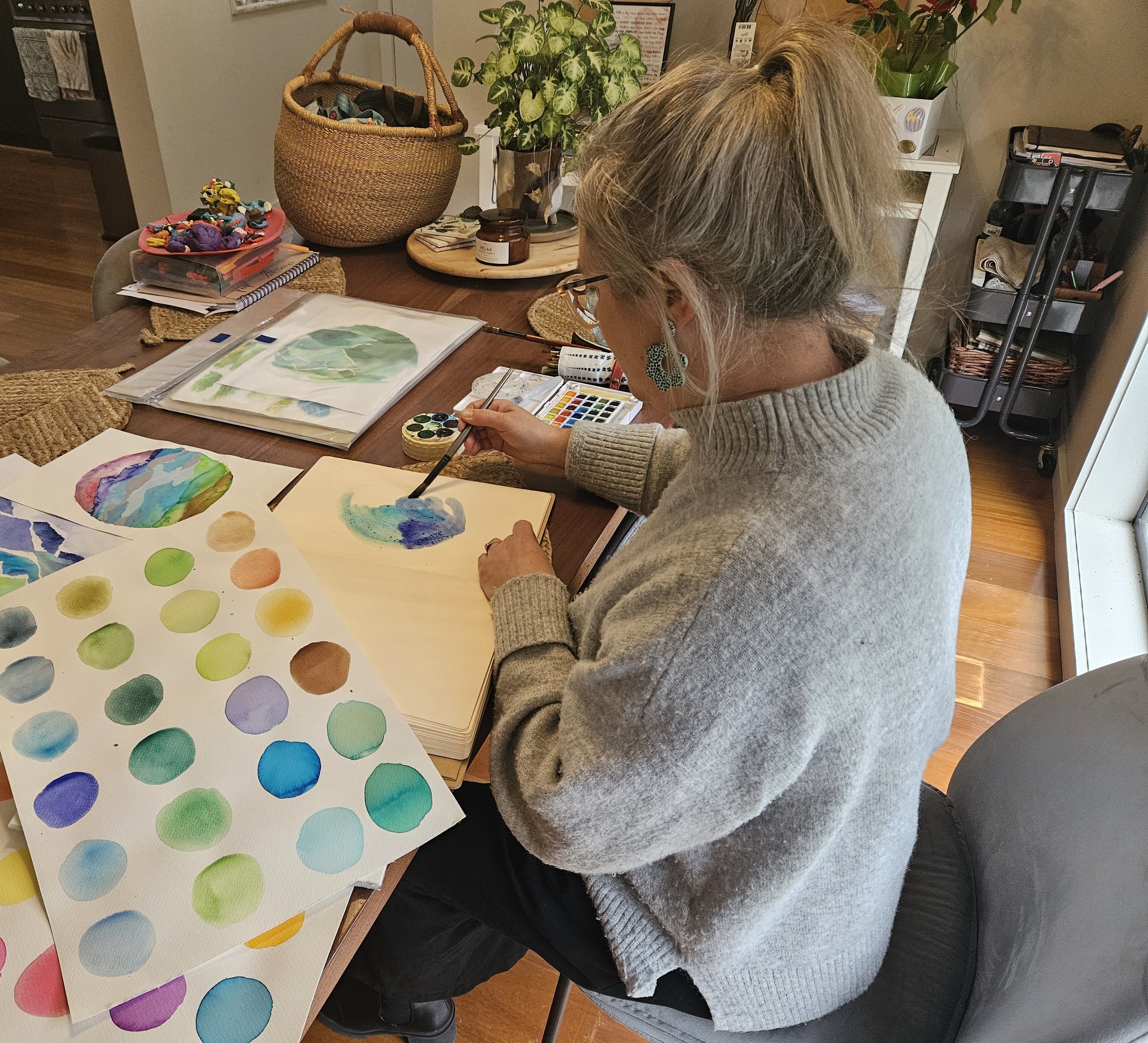 A woman with gray hair and glasses is sitting at a table, painting watercolor circles on paper. The table has watercolor paintings, a watercolor palette, and an art supply organizer. In the background, there are plants, a basket, and a framed piece of art.