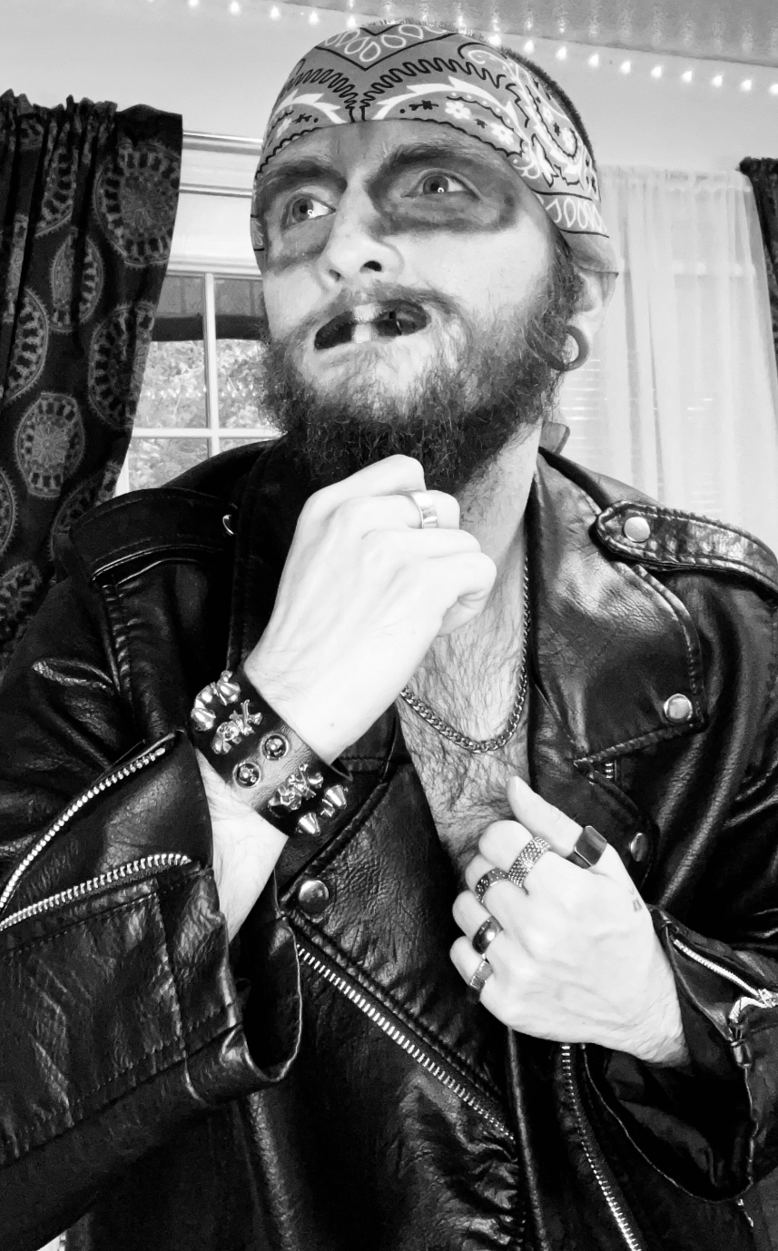 A man with a beard and mustache, wearing a bandana and leather jacket, makes a contemplative gesture with his hand near his chin. The photo is in black and white.
