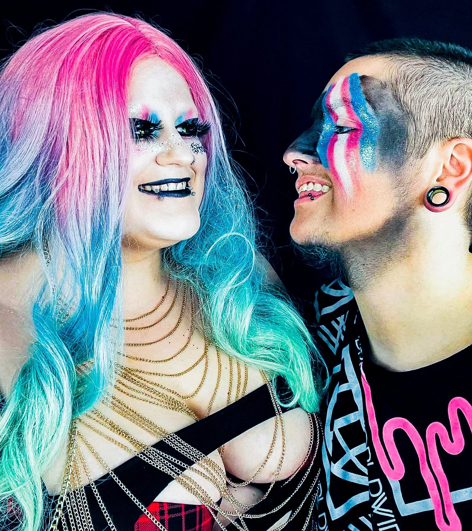 Two people with colorful and elaborate makeup and hair facing each other, smiling. The person on the left has pink, blue, and white hair, gold jewelry, and piercings. The person on the right has colorful face paint, a shaved hairstyle, and gauges.