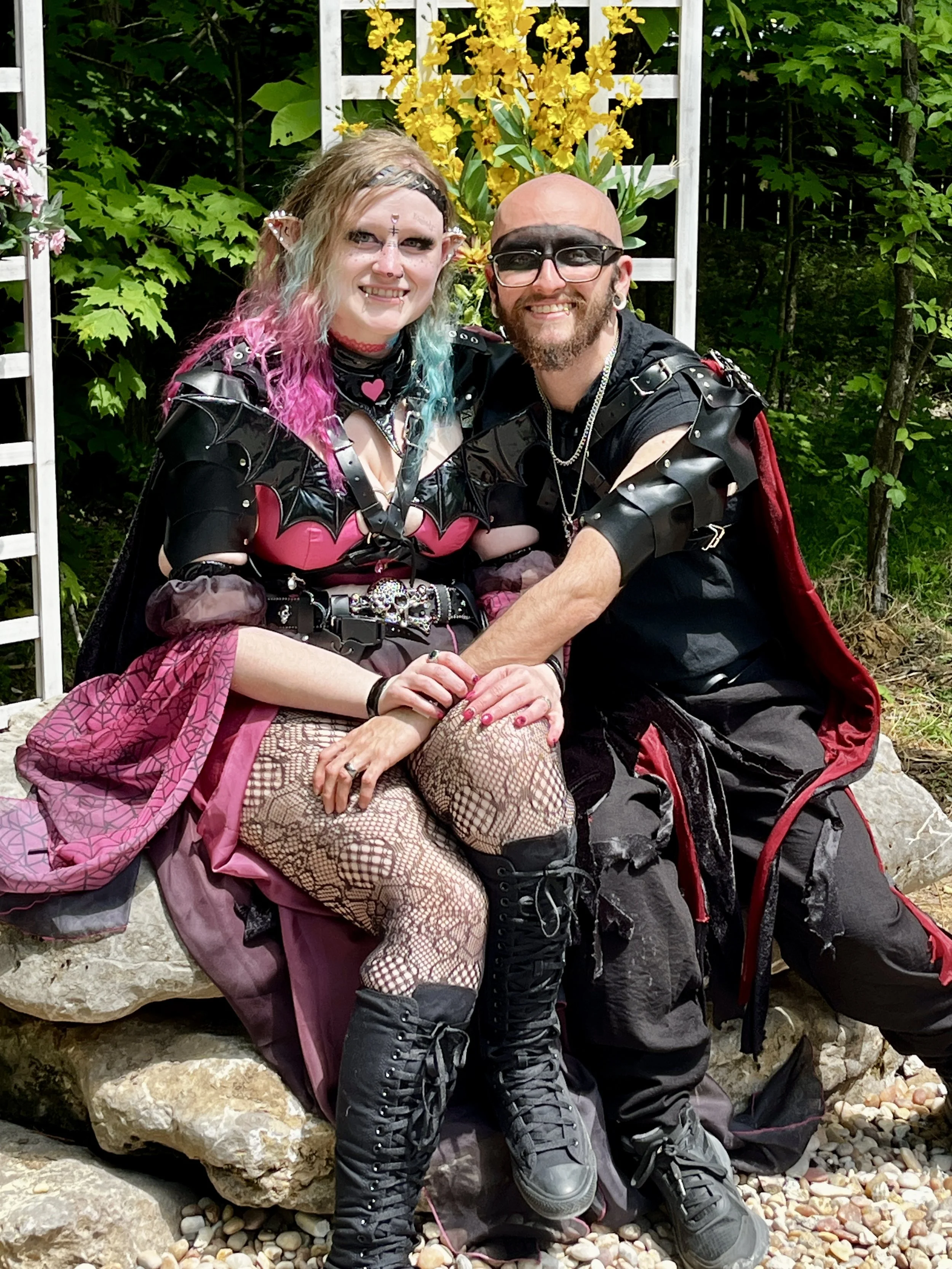 A smiling couple dressed in goth and punk fashion, sitting outdoors on rocks with trees and yellow flowers in the background.