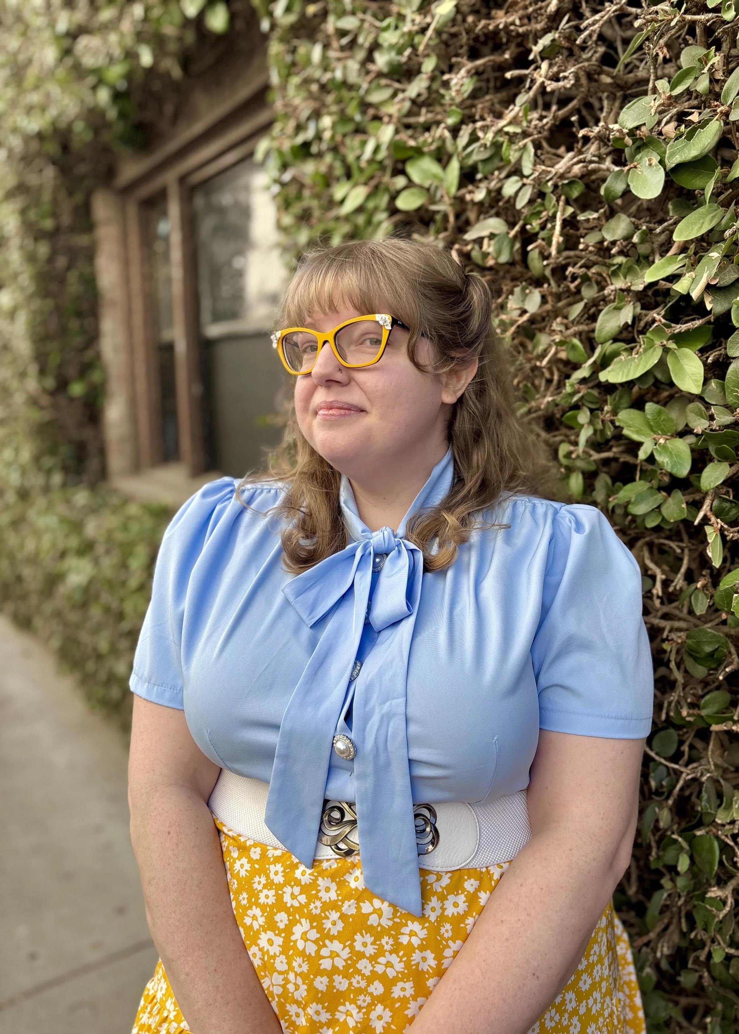 Woman with dark blonde, curly hair, wearing yellow glasses with decorative stones, a blue blouse with a bow, and a yellow skirt with white daisies, standing in front of greenery and a window.