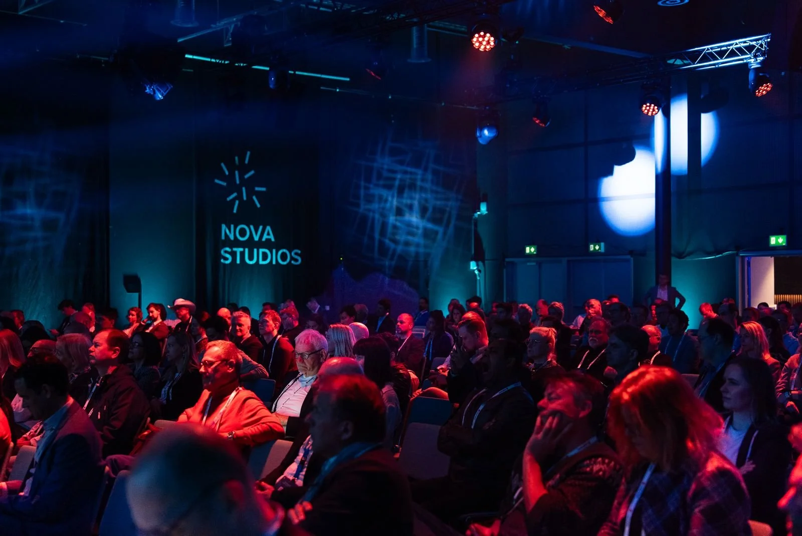 Audience attending a conference or event in a dimly lit room with a screen displaying 'NOVA STUDIOS' logo on the stage.