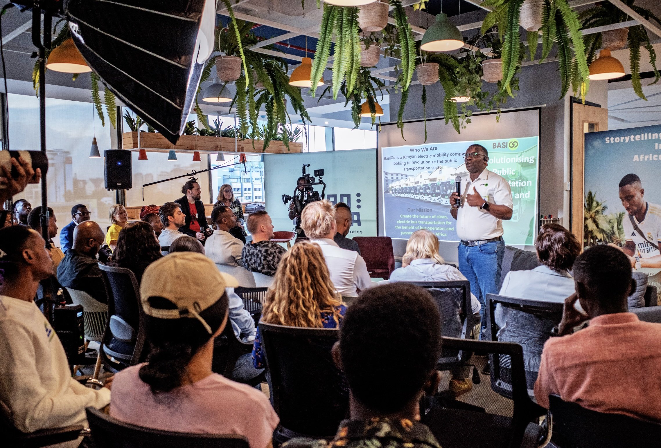 A man is giving a presentation on stage to an audience in a modern, well-lit room with hanging plants, large windows, and a large screen behind him displaying information about a Kenyan electric mobility company called BasiCo.