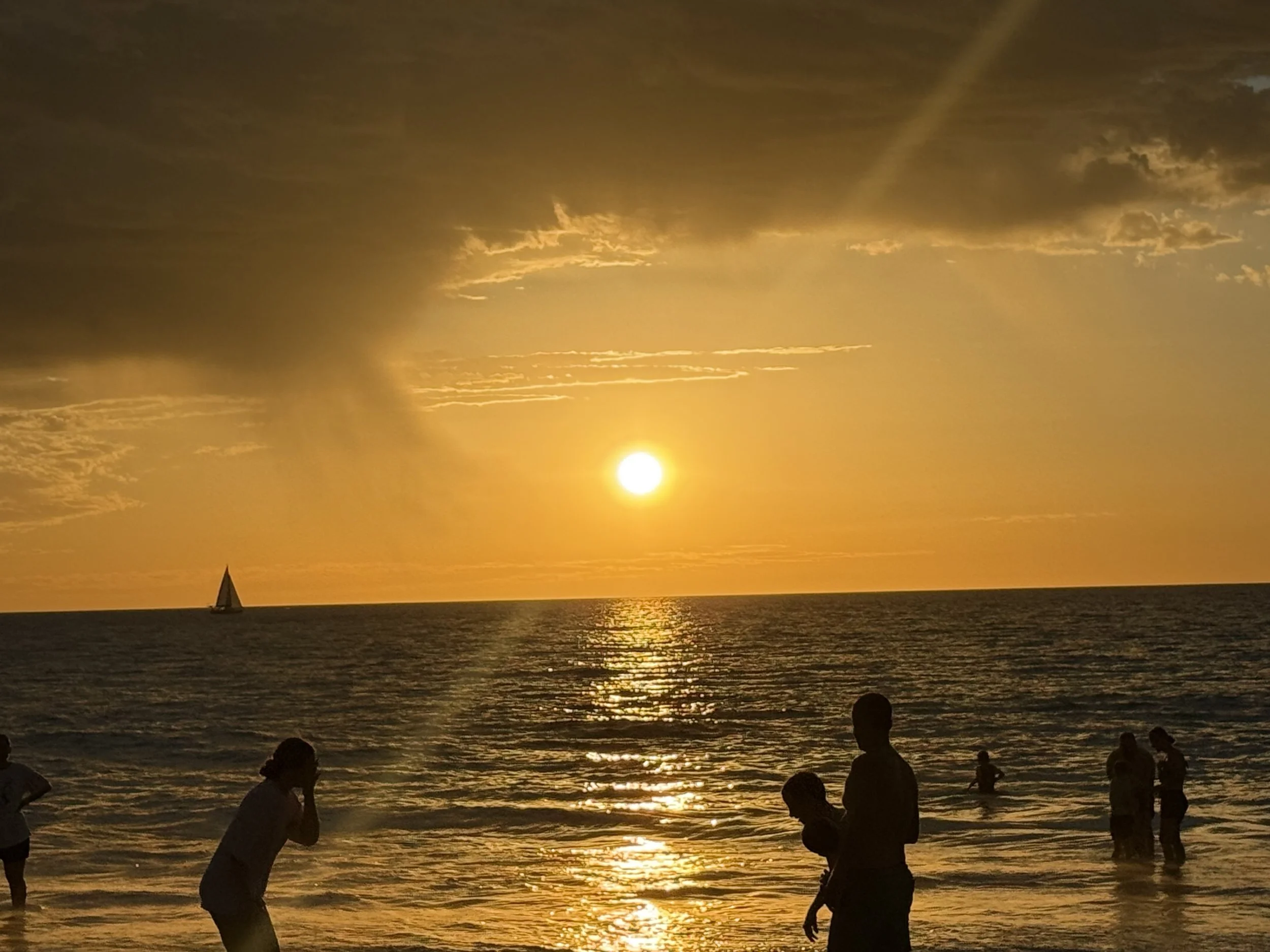 Sunset over the ocean with clouds in the sky. Sailboat visible on the water. People on the beach silhouetted against the sunset.