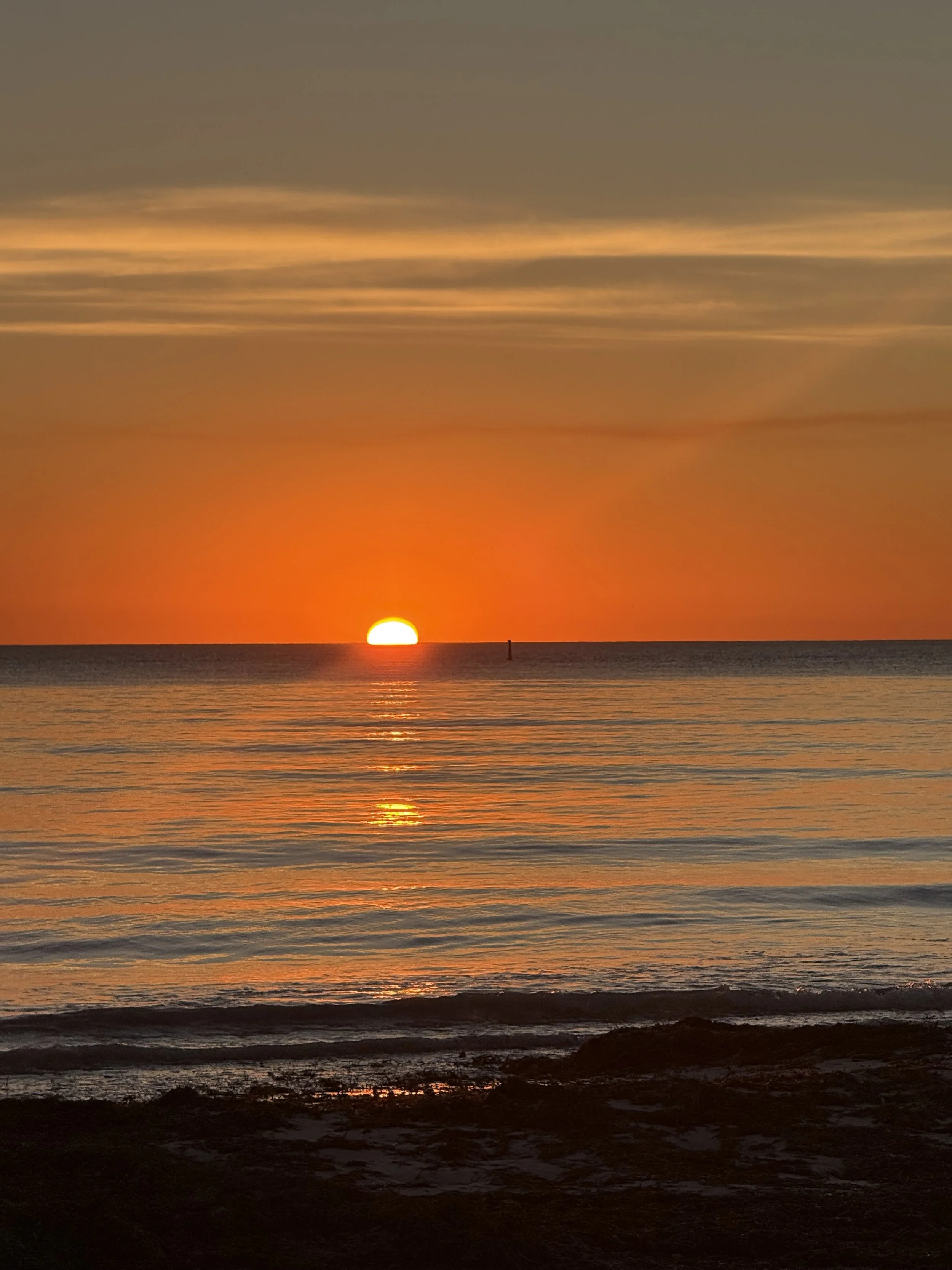 A sunset over the ocean with the sun partially below the horizon, reflecting orange and yellow hues on the water, and a dark rocky shoreline in the foreground.