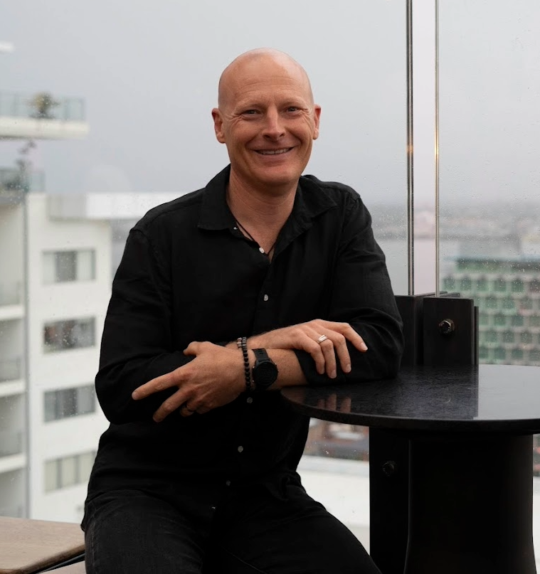 Smiling bald man in black shirt sitting by a window with an urban cityscape in the background.