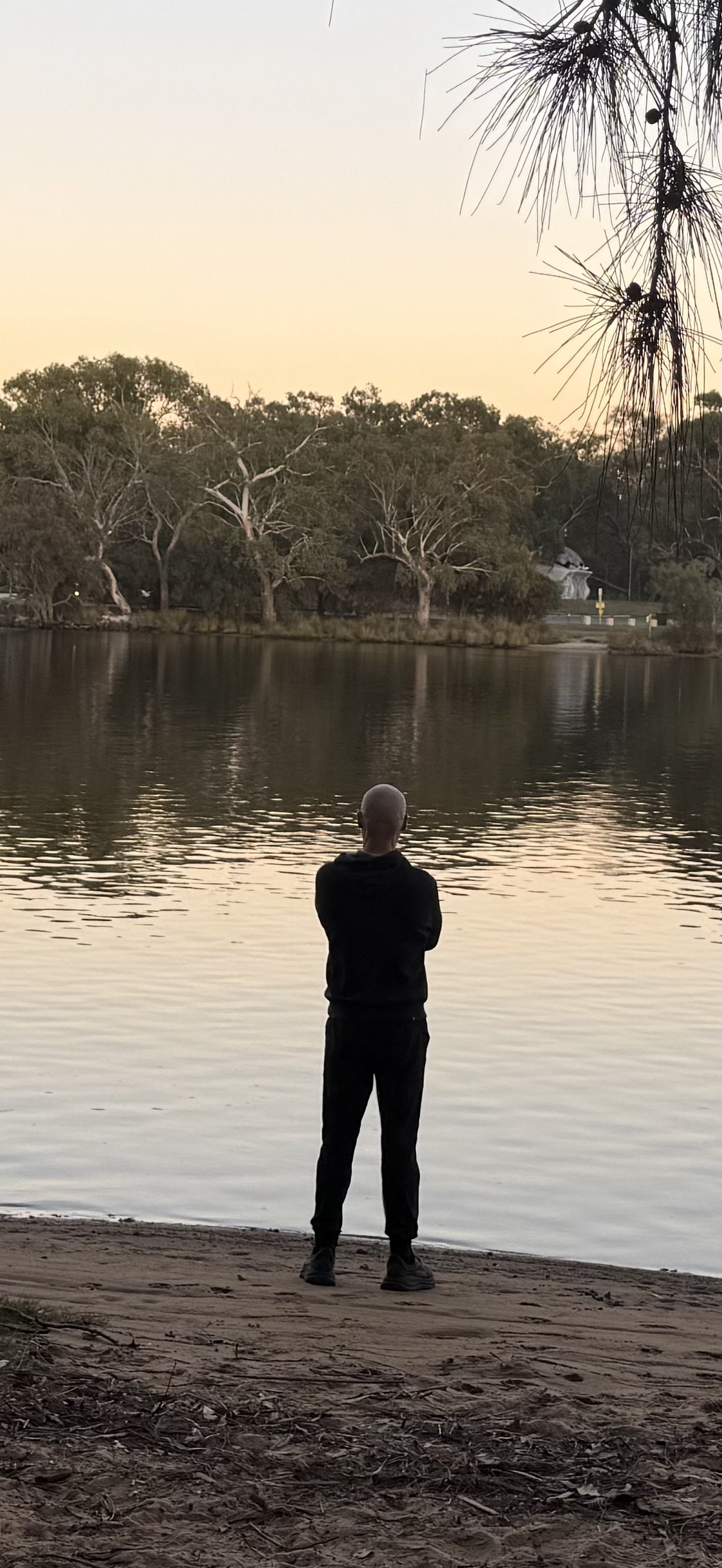 A person stands on the sandy shoreline of a body of water, facing away with arms crossed, during sunset. The background shows trees across the water and a pale sky.