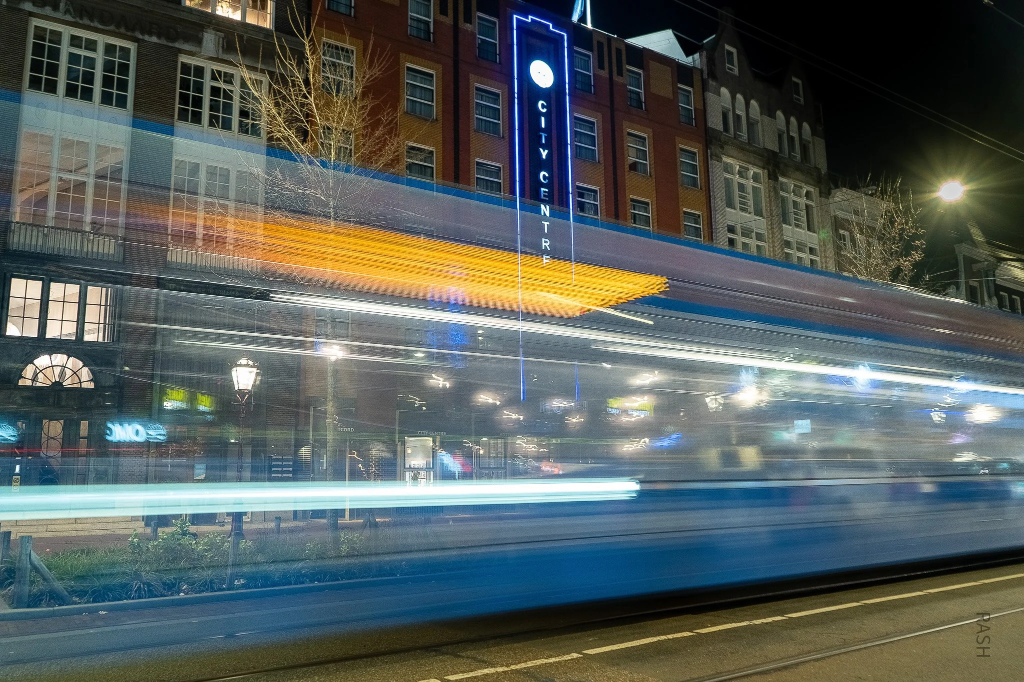 Nighttime city street scene with a building featuring a vertical sign reading 'City Centre' in blue neon lights, light trails from moving vehicles, and illuminated storefronts at the ground level.