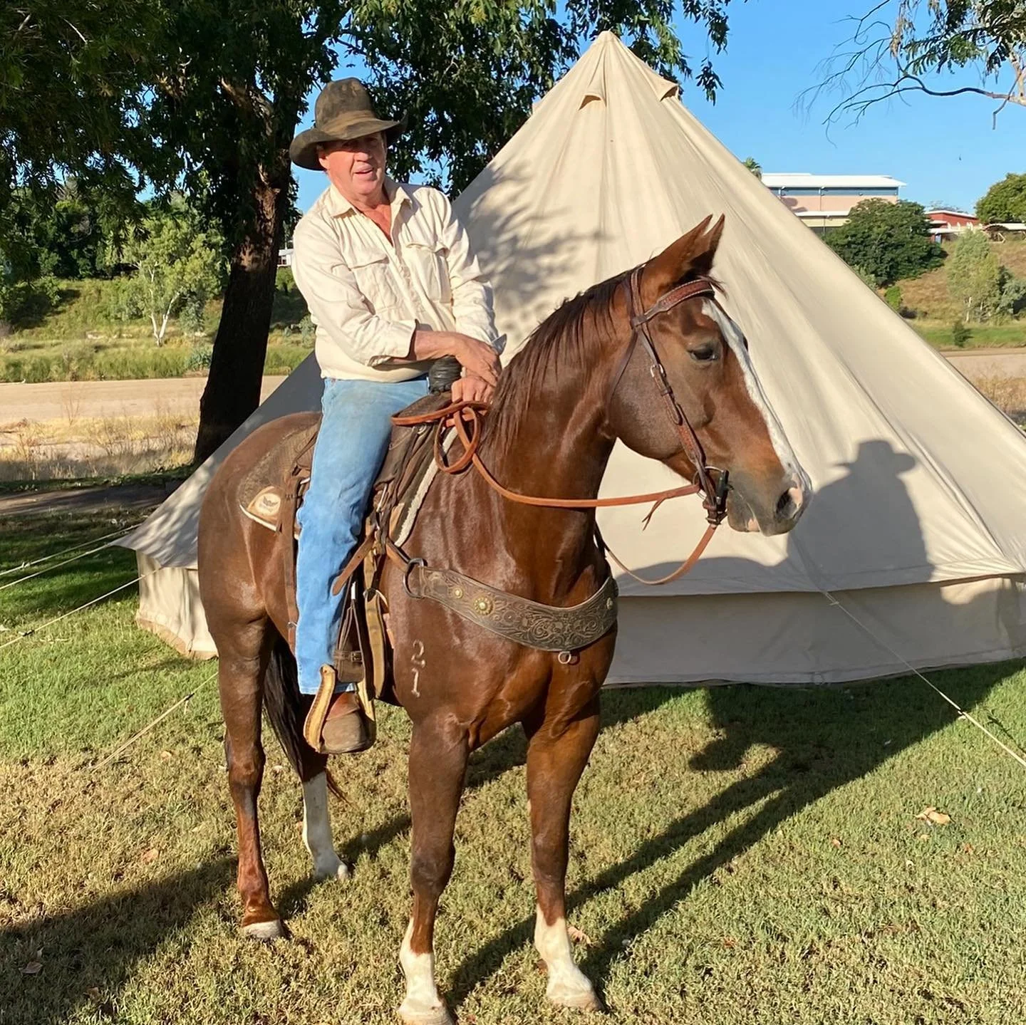 A man wearing a cowboy hat and light-colored shirt riding a brown horse with a white blaze on its face, in front of a large beige teepee, with trees and a building in the background on a sunny day.